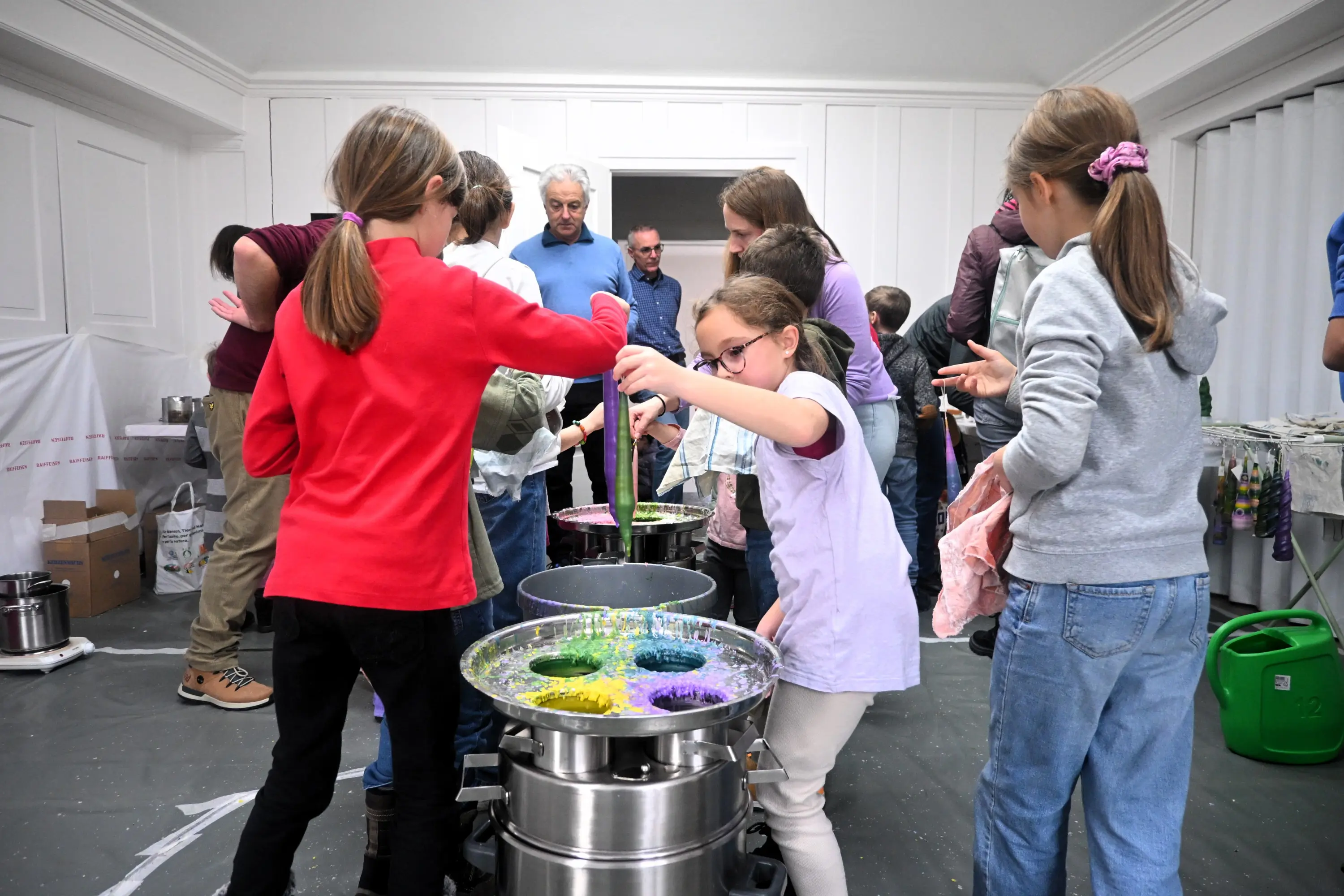 Bambini mescolano coloranti vivaci in grandi contenitori durante un'attività di arte creativa. Alcuni adulti osservano da dietro. L'ambiente è luminoso e
