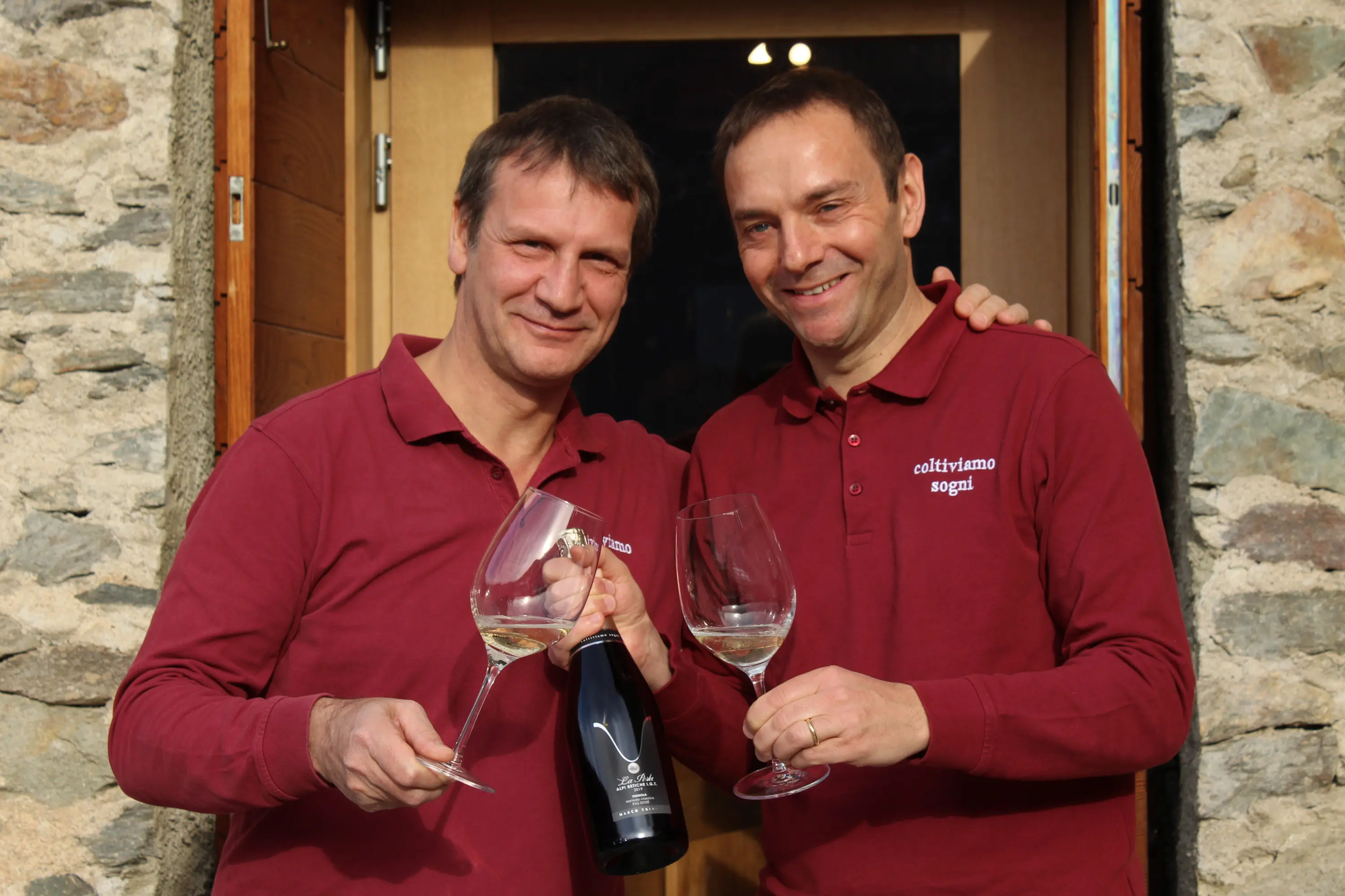 Due uomini sorridenti si trovano davanti a una porta in legno, entrambi indossano magliette rosse. Uno tiene una bottiglia di vino e l'altro un calice