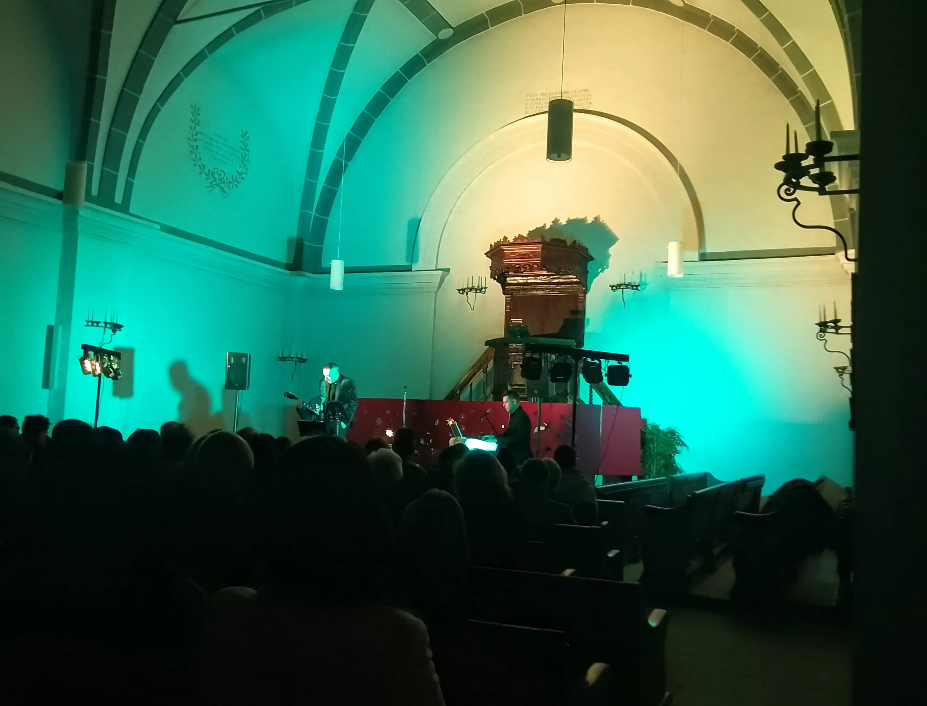 Un concerto si svolge in una chiesa con illuminazione verde e blu. Un musicista è sul palco con un altro membro del gruppo che suona a fianco. Il pubblico è visibile