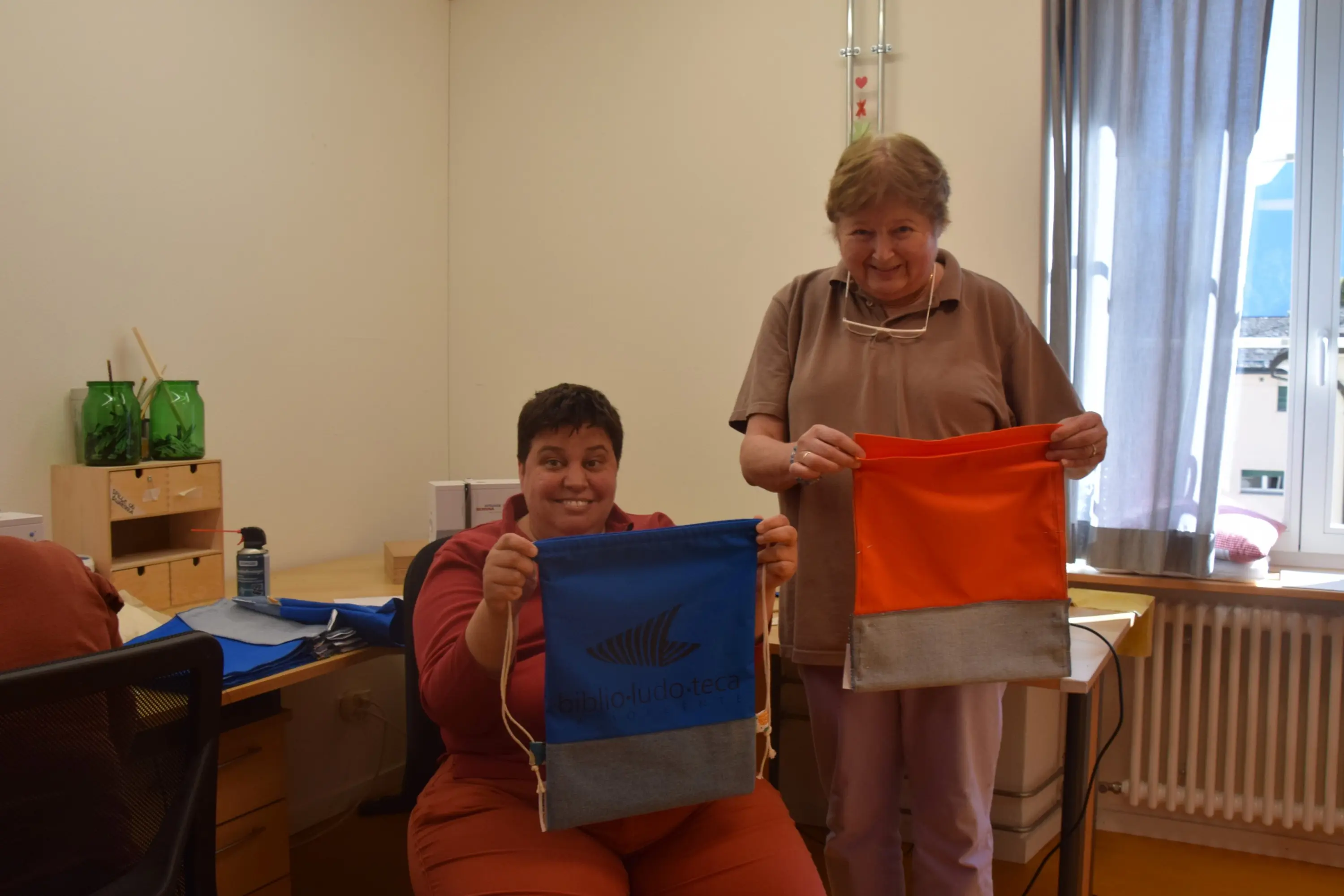 Due donne sorridenti mostrano delle borse colorate. Una tiene una borsa blu con il logo della biblioteca, mentre l'altra tiene una borsa arancione. Sullo sfondo si