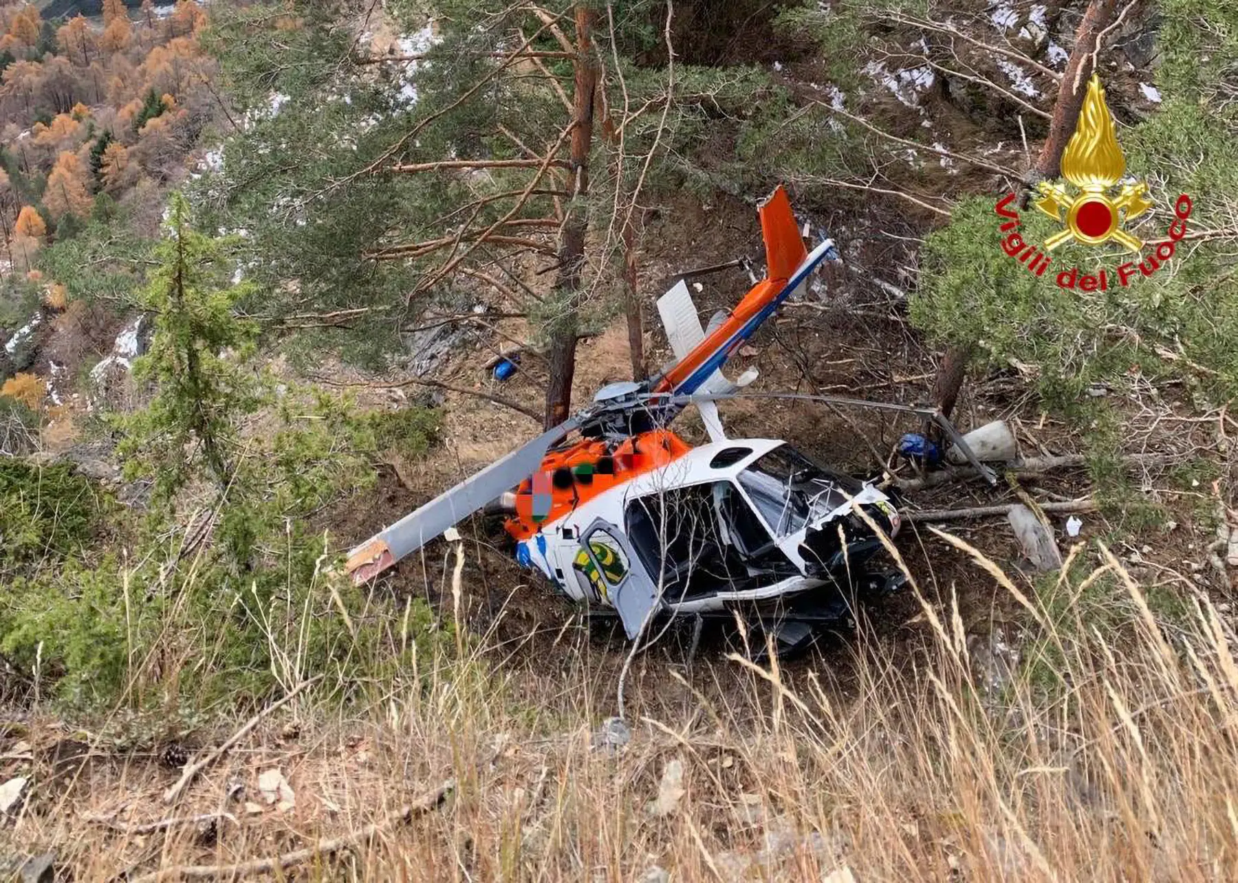 Un elicottero schiantato tra gli alberi in un'area montuosa, con vegetazione circostante e detriti visibili.