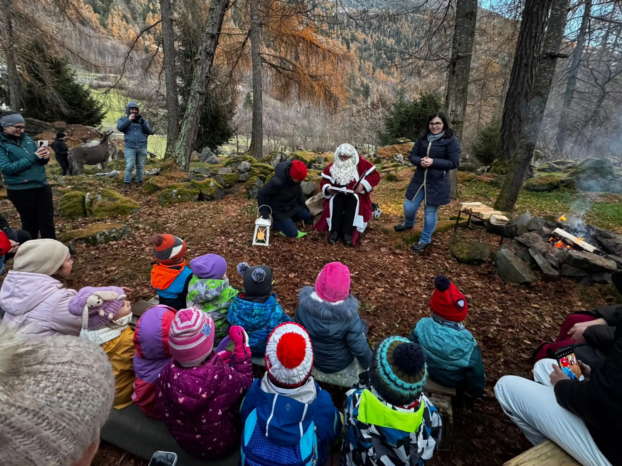 Babbo Natale è seduto su un tronco, circondato da bambini vestiti con giacche colorate. Alcuni adulti assistono, mentre la scena si svolge in un