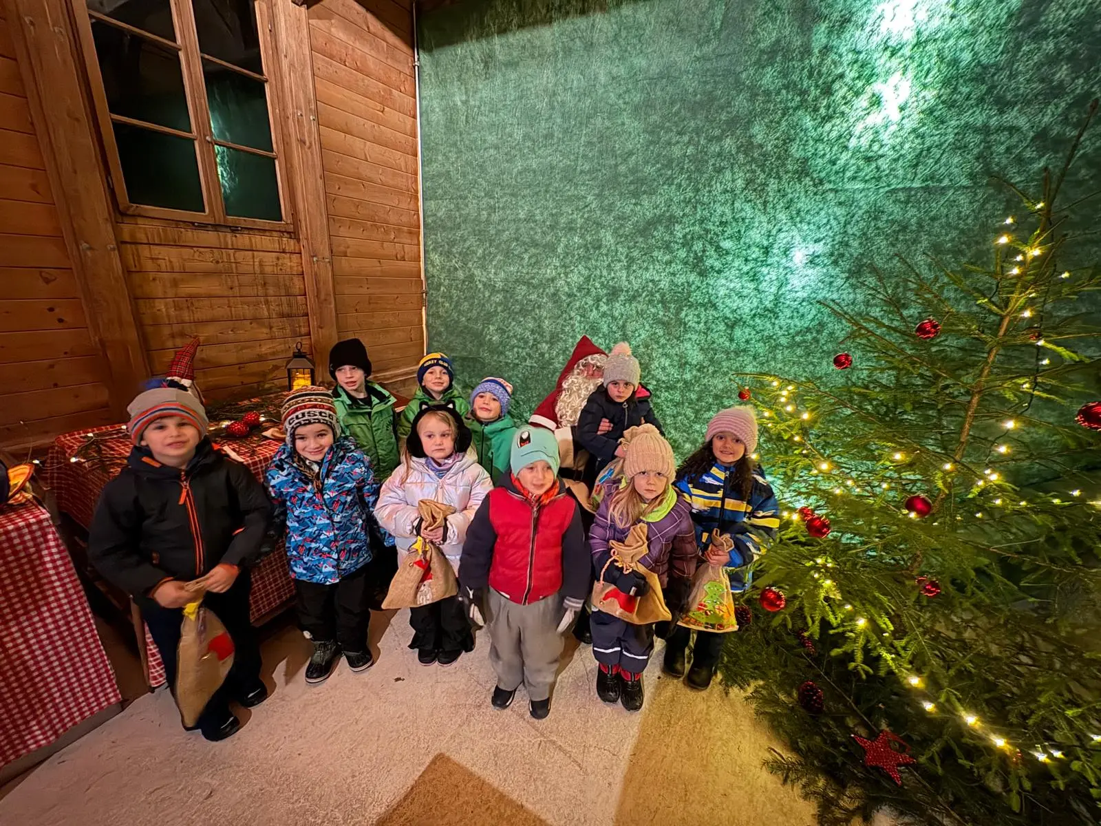 Un gruppo di otto bambini, vestiti con giacche invernali e berretti, sorridono davanti a un grande albero di Natale decorato con palline rosse, in