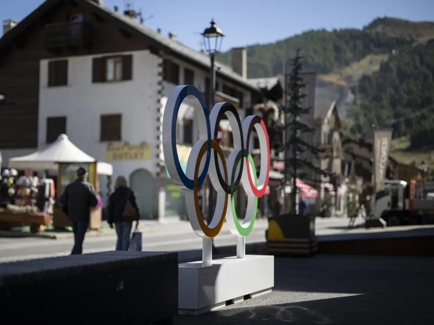 Cinque cerchi olimpici in colore blu, giallo, nero, verde e rosso, esposti in una piazza con edifici tipici di montagna sullo sfondo.