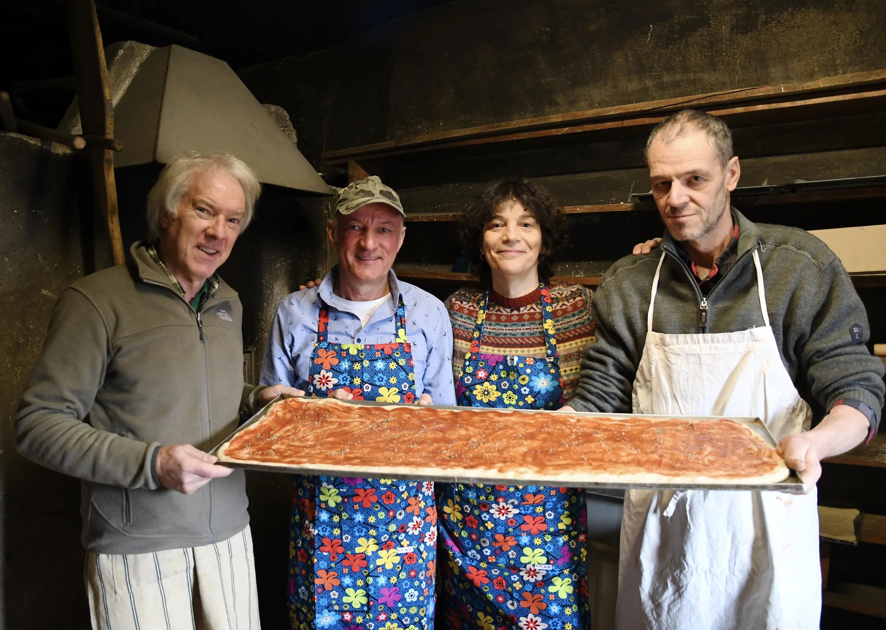 Quattro persone sorridenti mostrano una teglia con un impasto per pizza. Indossano grembiuli colorati e si trovano in una cucina rustica.