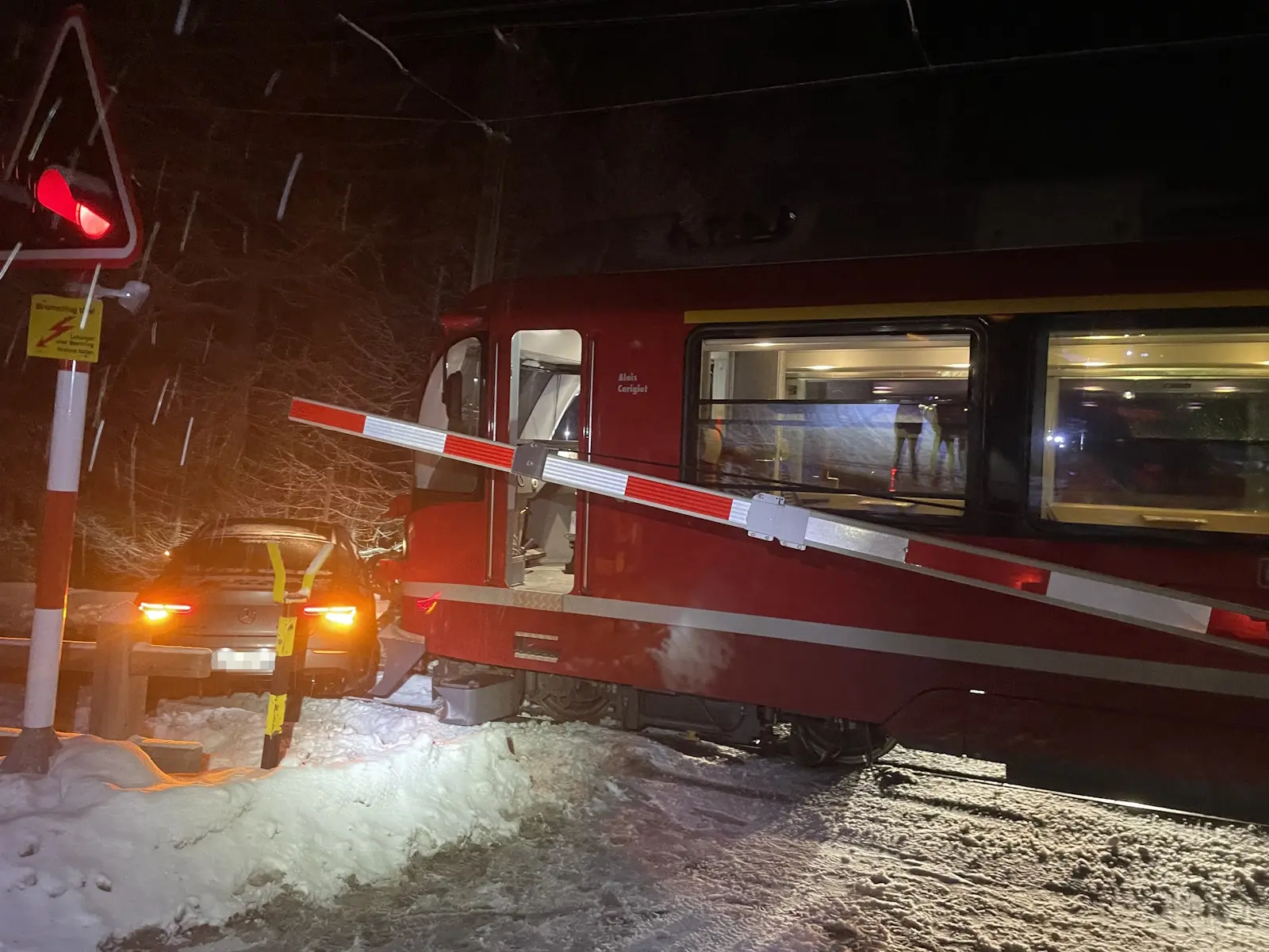 Treno rosso fermo vicino a un passaggio a livello con una sbarra abbassata, mentre un'auto si trova bloccata sulla neve. Neve che cade e oscurità