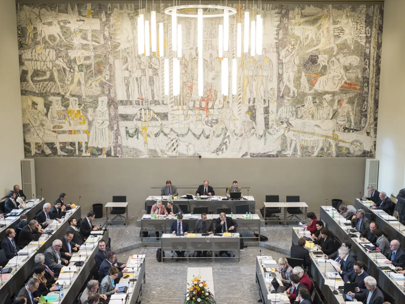 Un'assemblea con molti membri seduti in una sala moderna, con un grande murale decorativo sulla parete. Ci sono tavoli disposti a semicerchio, e diverse persone sembr