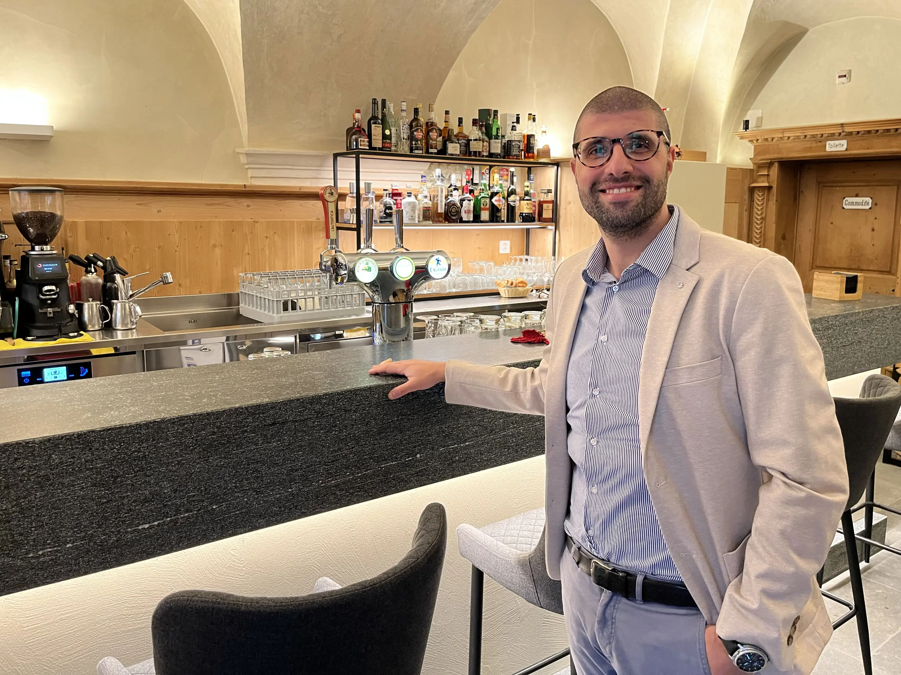 Uomo sorridente in giacca chiara accanto al bancone di un bar elegante, con bottiglie e attrezzature per il caffè sullo sfondo.