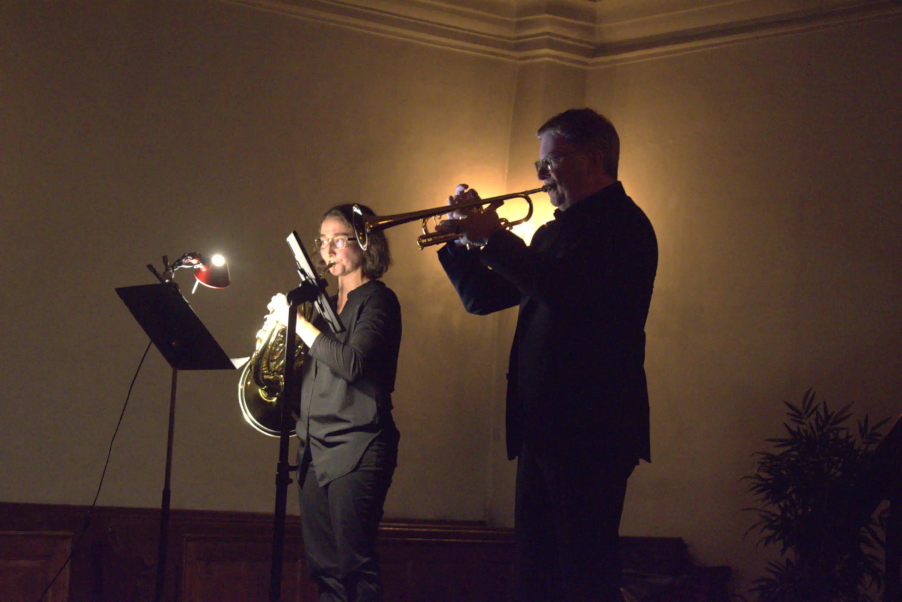 Un trombettista suona mentre una musicista suona l'eufonio. Entrambi sono in abiti neri e sono illuminati da una luce soffusa sullo sfondo chi