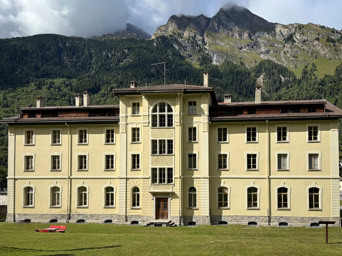 Edificio di colore giallo con finestre a bovindo, situato in un'area montuosa. Sullo sfondo si vedono montagne coperte di vegetazione e nu