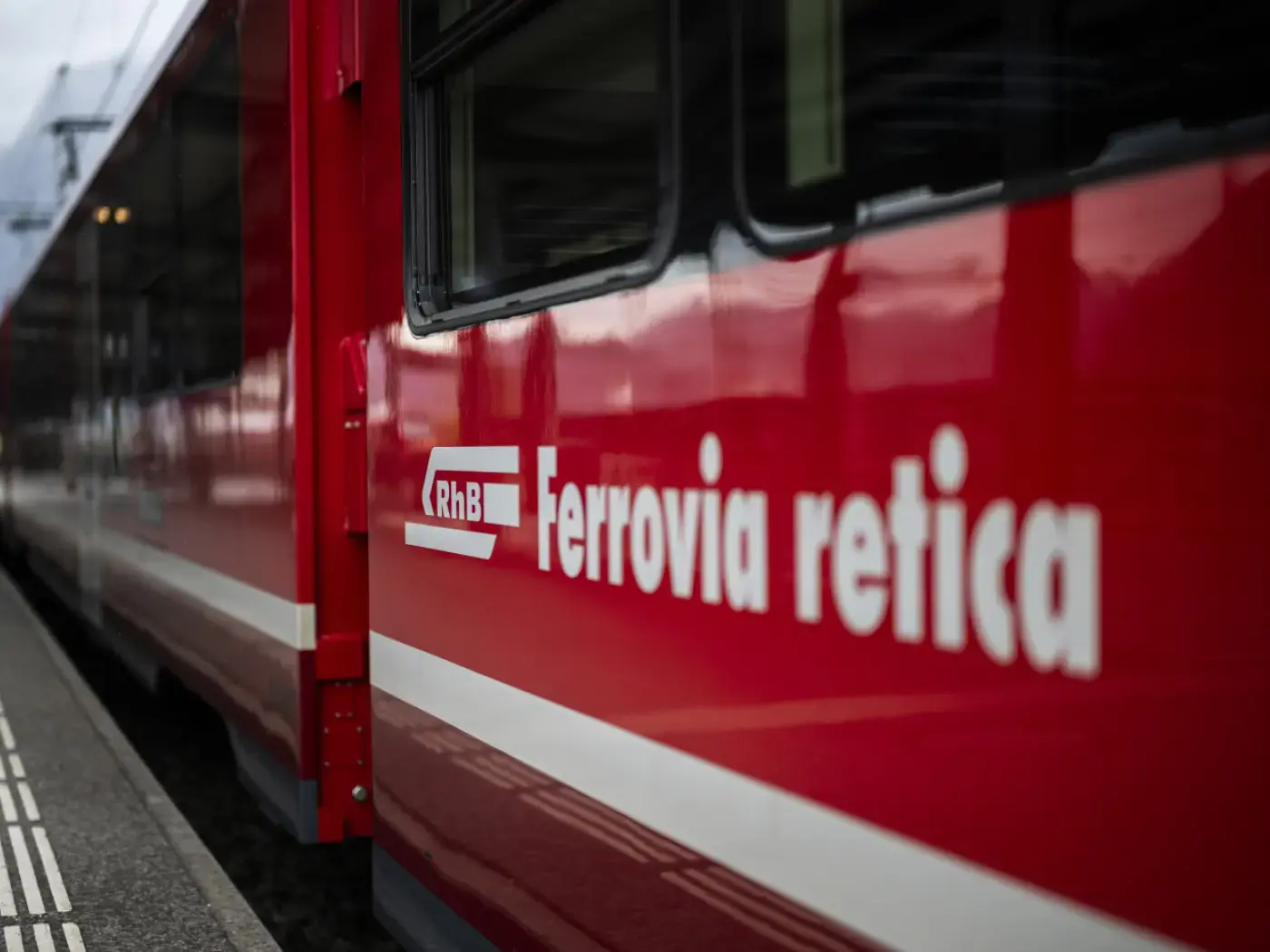 Treno rosso della Ferrovia Retica, con logo visibile e riflessi sul vetro. Atmosfera urbana e ferroviaria.