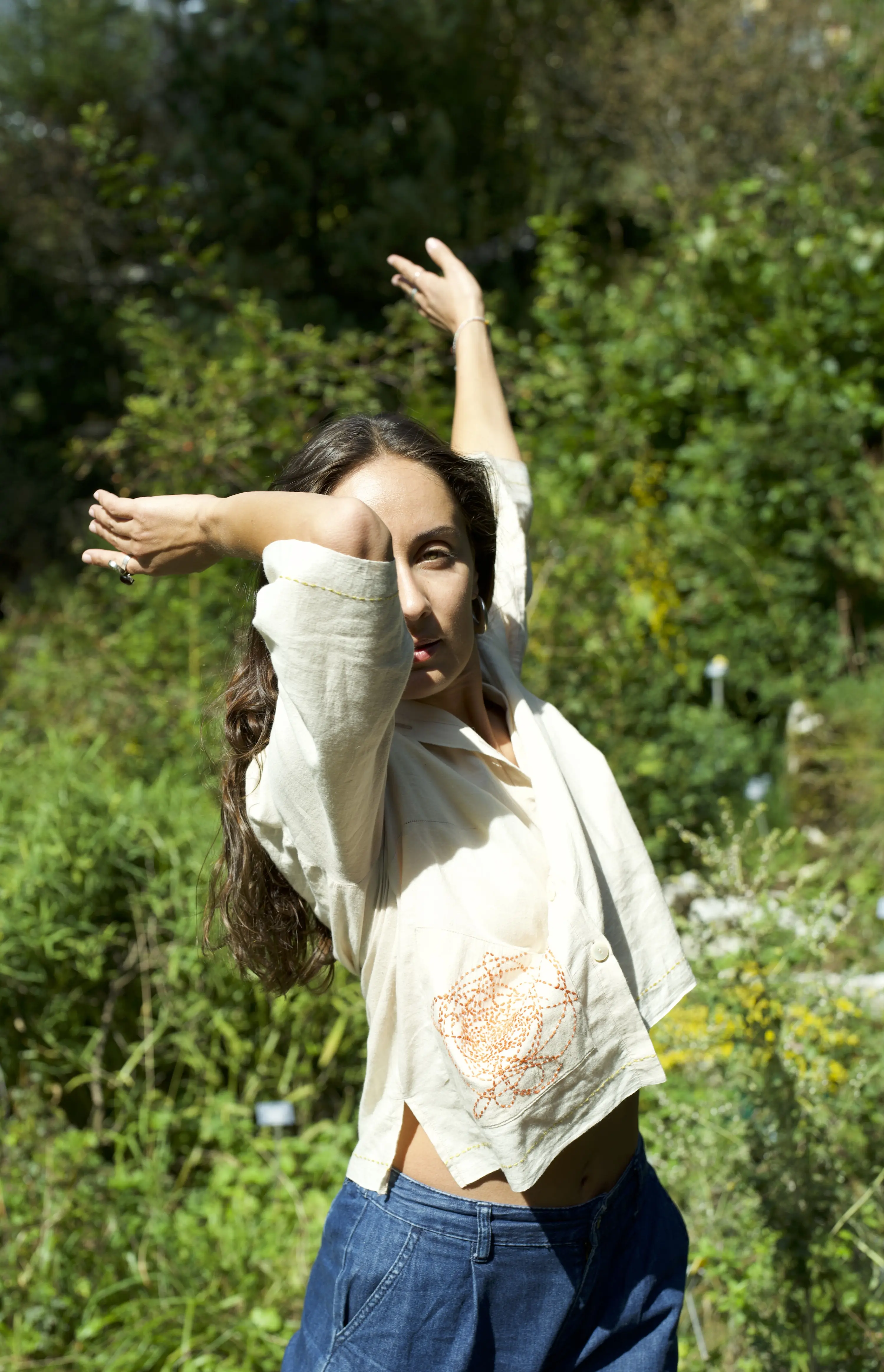 Donna con lunghi capelli mossi, che indossa una camicia beige con ricamo, in posizione di danza all'aperto, circondata da vegetazione.