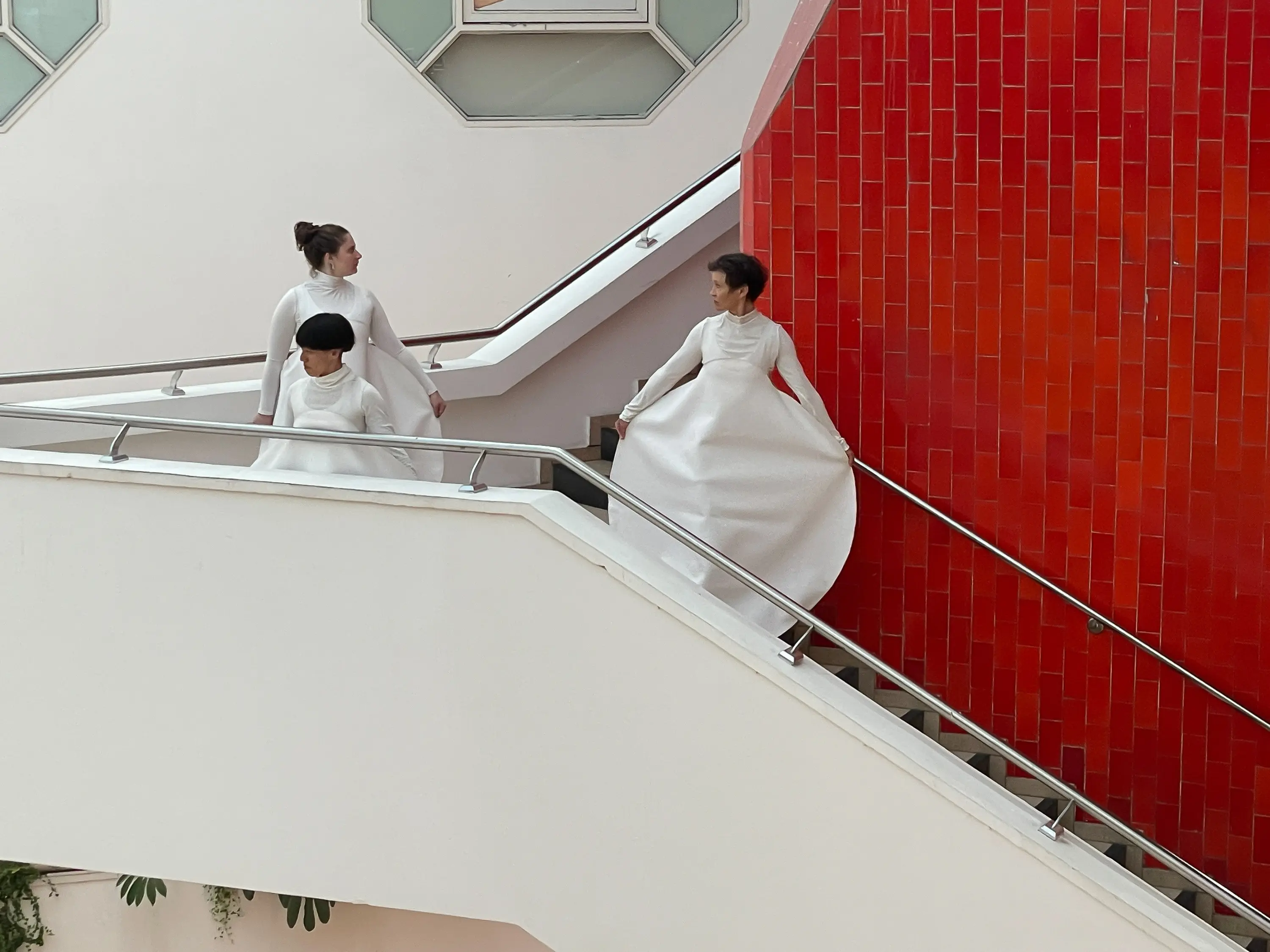 Tre donne in abiti bianchi scendono una scala. Due di loro indossano gonne ampie che fluttuano mentre si muovono, mentre una terza le osserva.