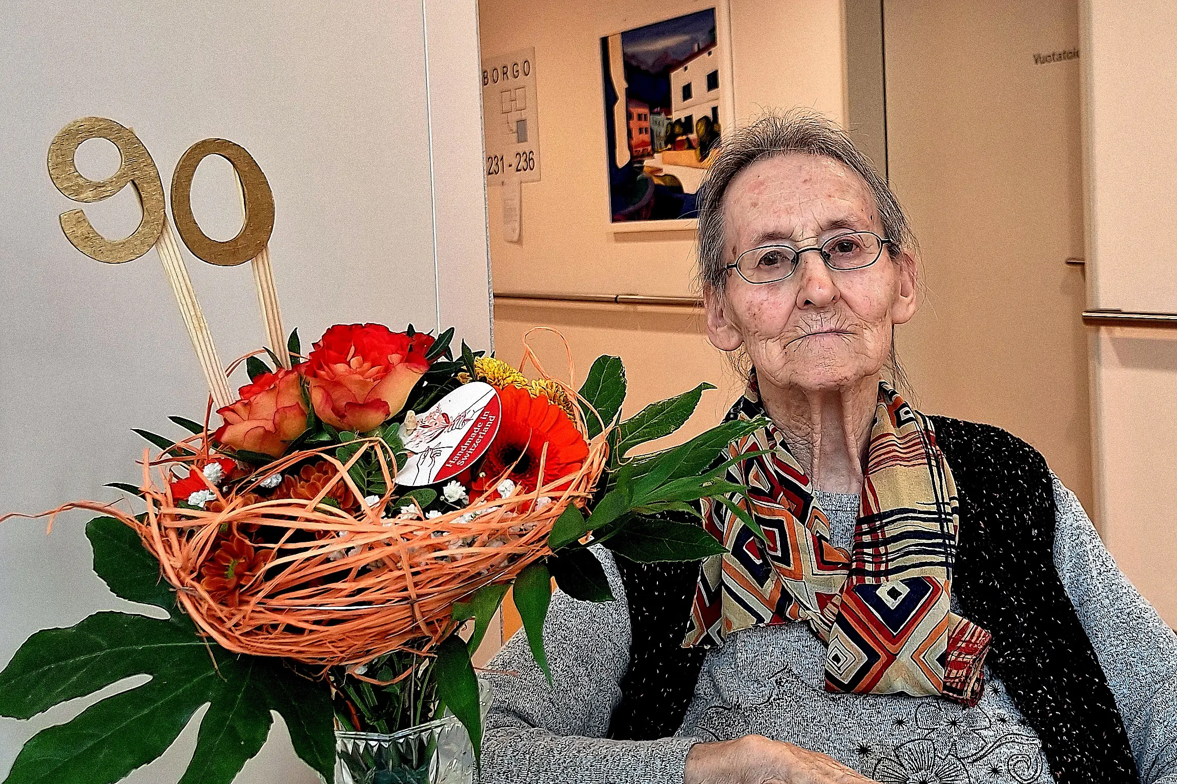 Una donna anziana, seduta, con un grande bouquet di fiori e un cartello che indica "90", in un ambiente caldo e festoso.