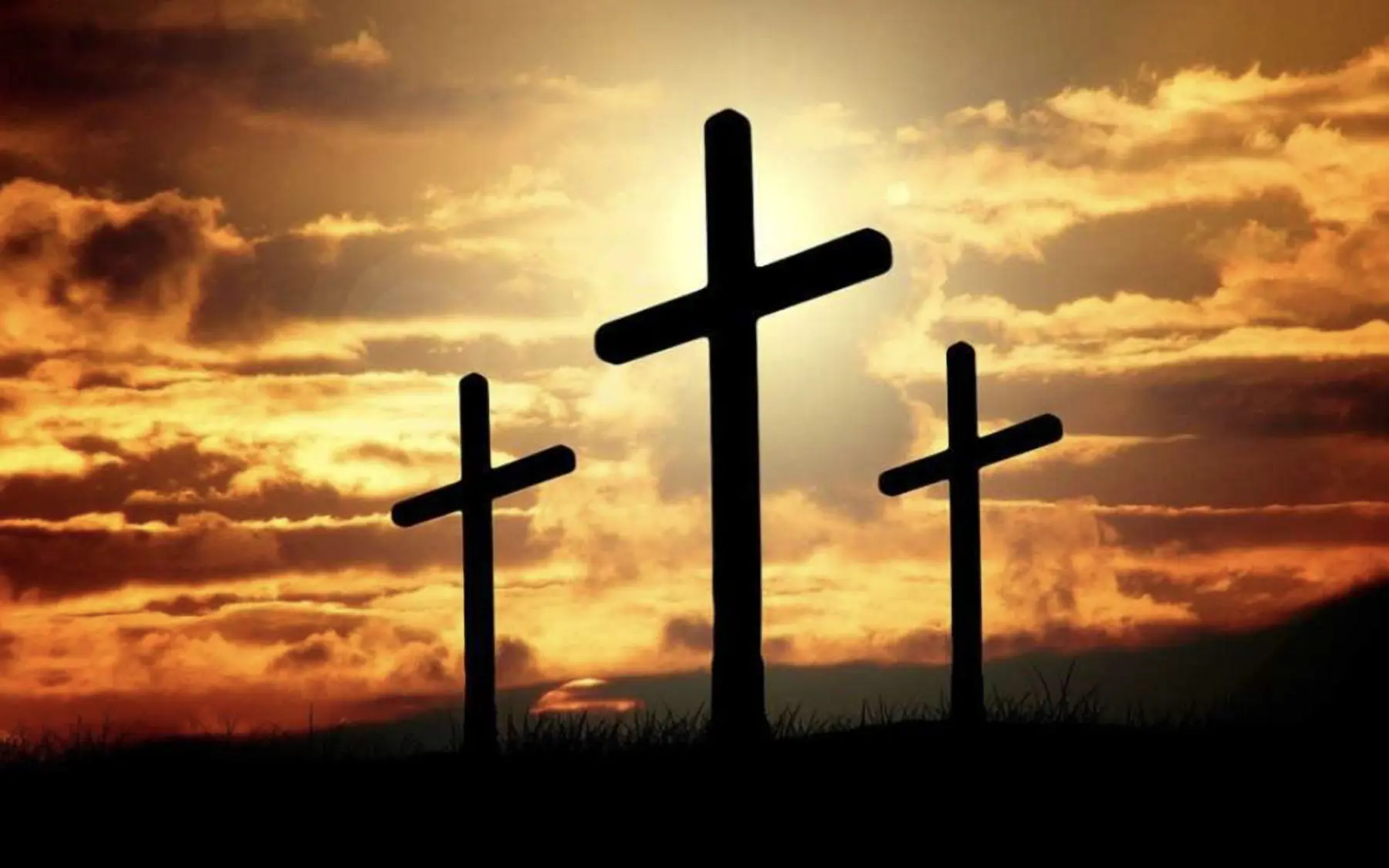 Tre croci scure si stagliano contro un cielo al tramonto, con nuvole illuminate da tonalità calde di arancione e giallo. La luce solare riflette