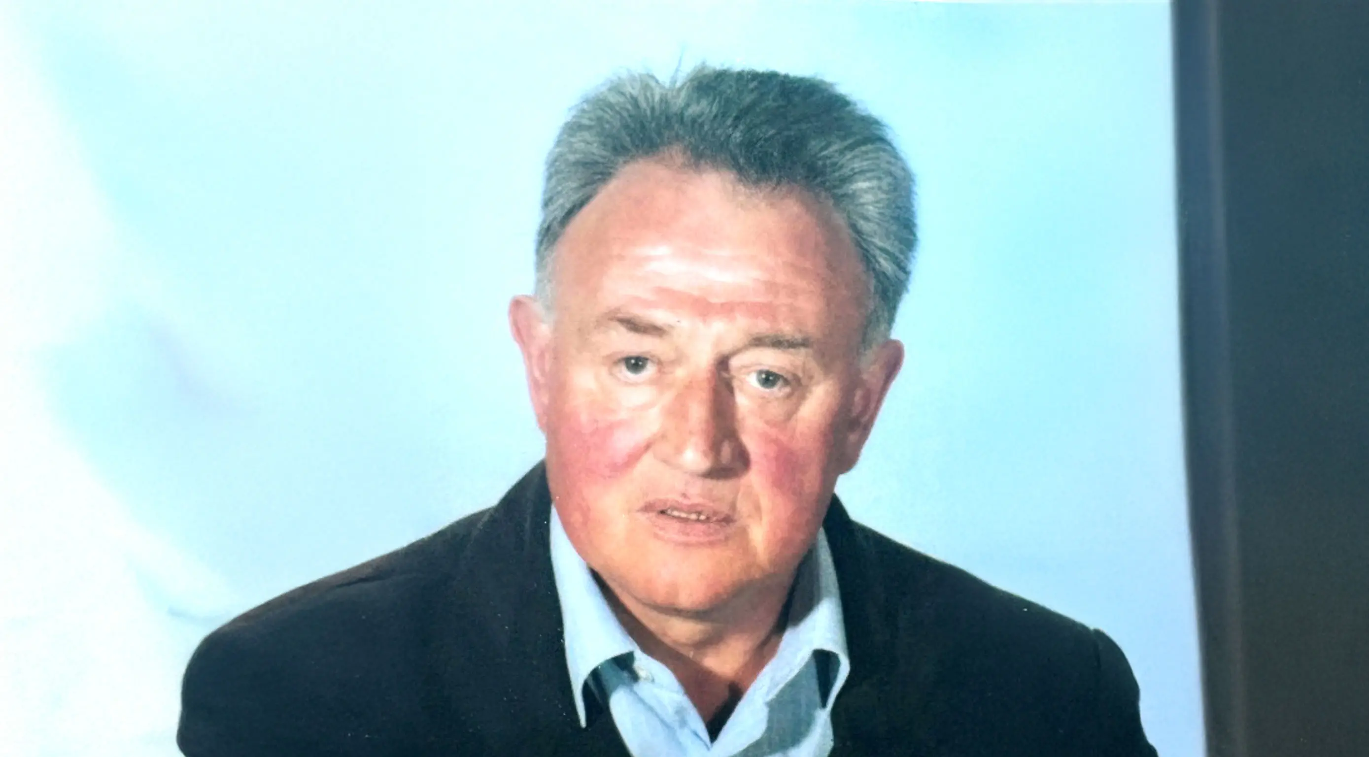 Un uomo anziano con capelli grigi, indossa una giacca scura e una camicia chiara. Ha un'espressione seria e guarda direttamente verso la fotocamera. Sullo sfondo c'è un'illuminazione morbida, con tonalità blu.