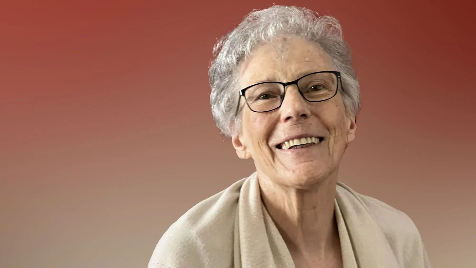 Una donna anziana con occhiali e capelli grigi sorridente. Indossa un maglione chiaro e si trova davanti a uno sfondo sfumato rosso.