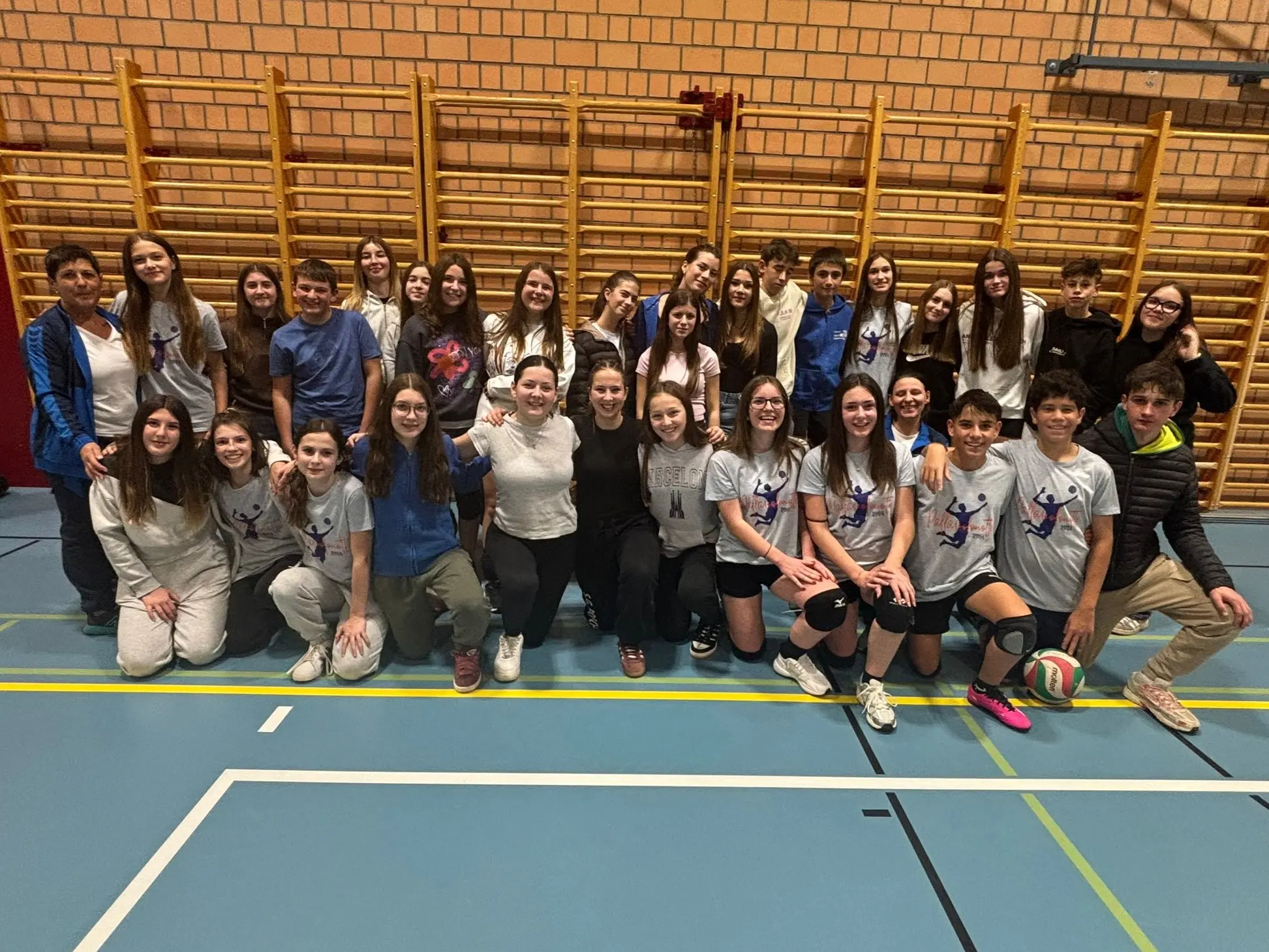 Gruppo di ragazzi e ragazze in una palestra, sorridenti e posati per una foto di squadra, con palloni da pallavolo e attrezzature sportive sul pavimento.