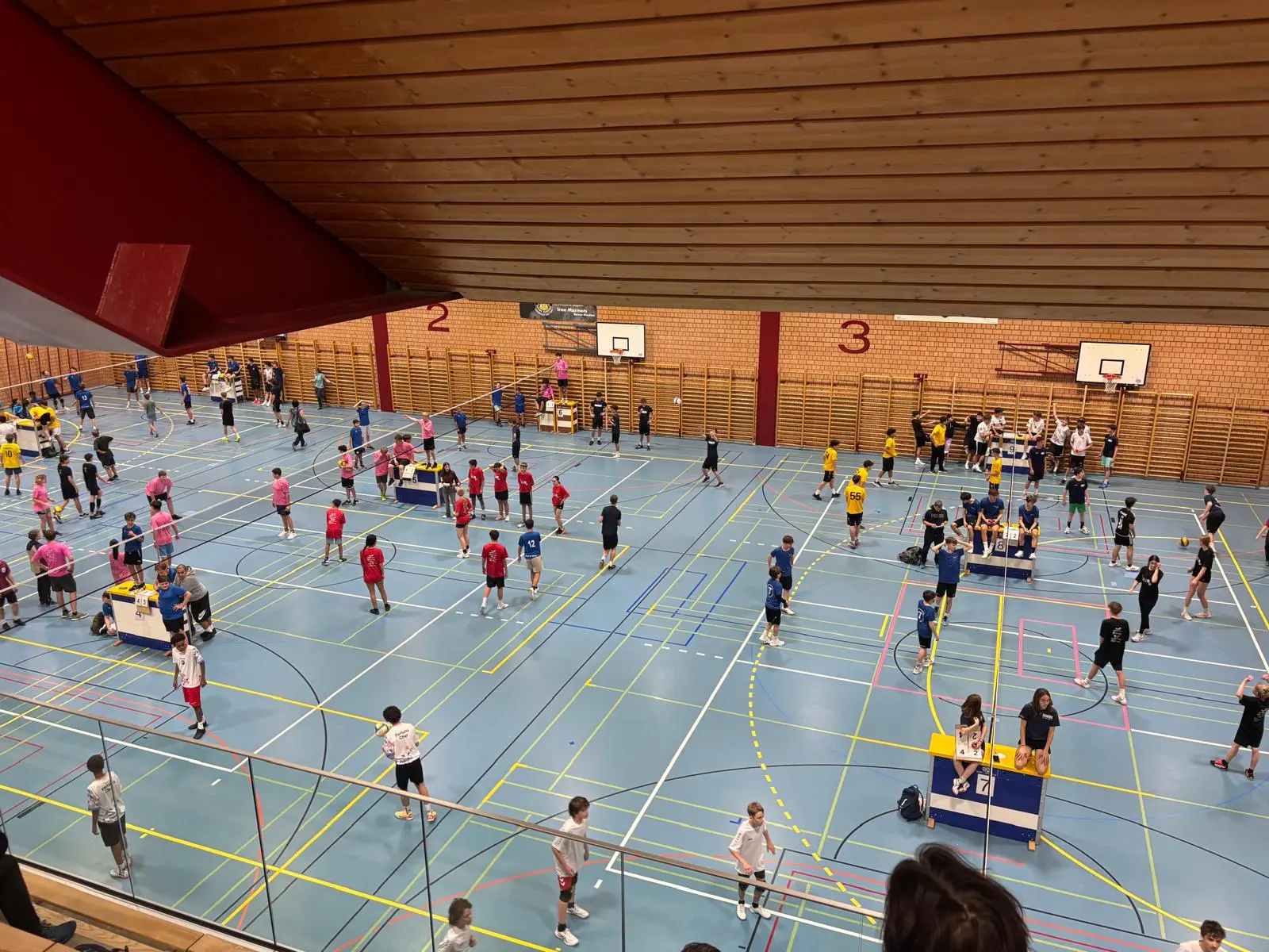 Una vista dall'alto di un campo sportivo, con diverse persone impegnate in attività. Ci sono gruppi di giovani vestiti con magliette di vari colori che si allenano o partecipano a una competizione. Il pavimento è segnato per diverse sport e ci sono can