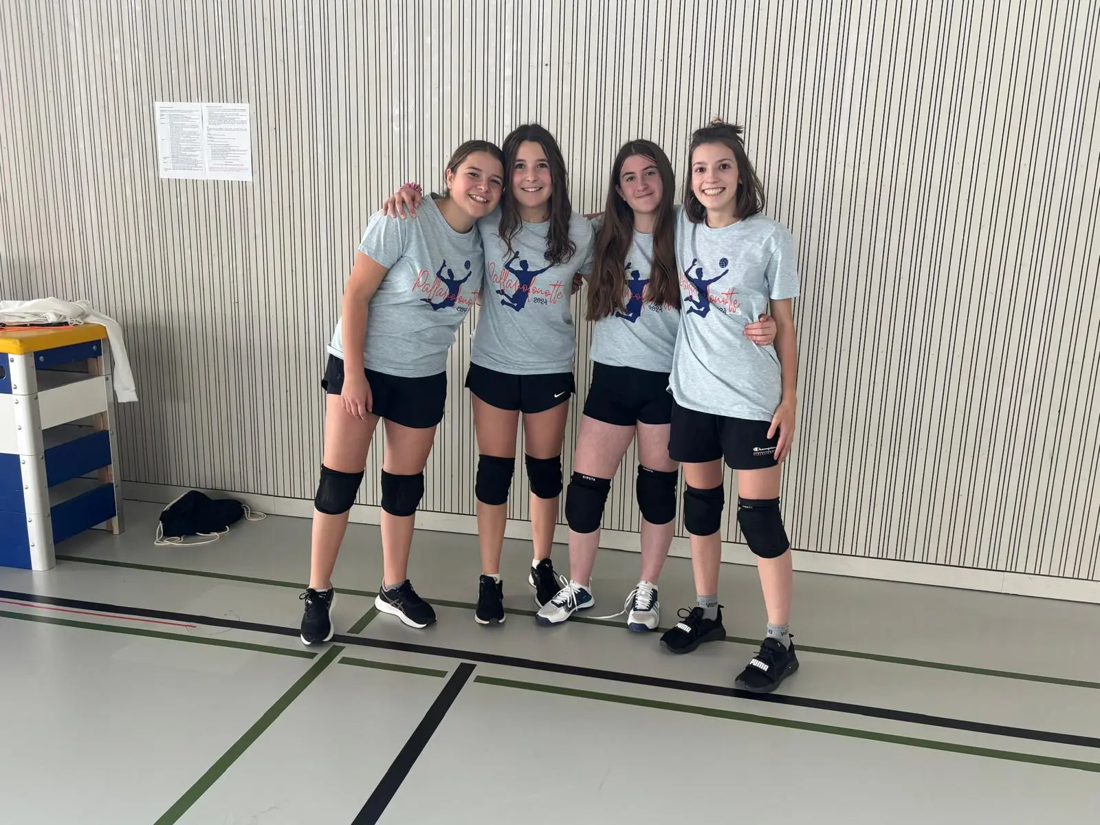 Quattro ragazze sorridenti in maglietta grigia e pantaloni corti, pronte per una partita di pallavolo. Indossano ginocchiere e si trovano in un palazzetto dello sport.