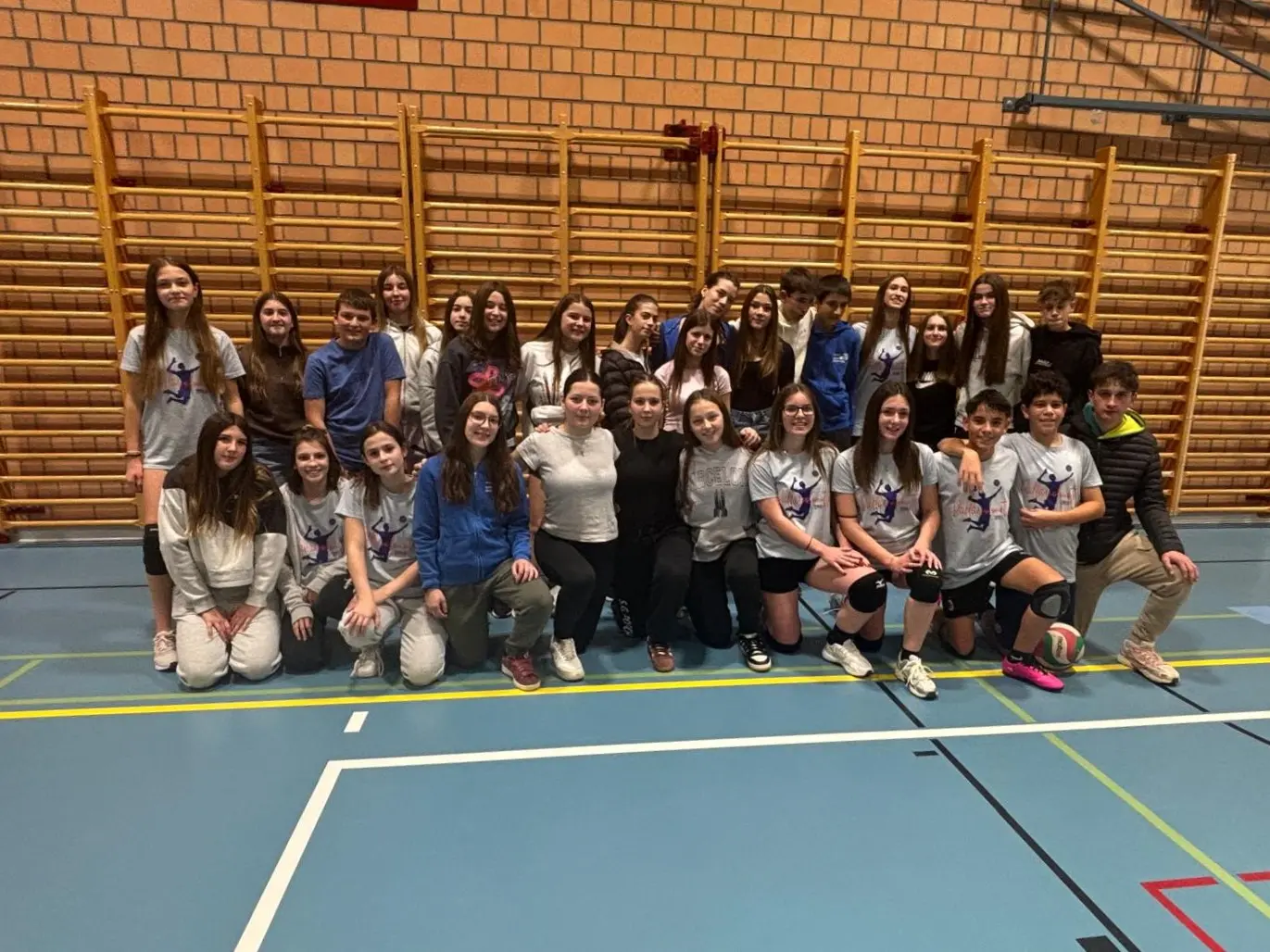 Gruppo di giovani atleti di pallavolo in una palestra, sorridenti e vestiti con magliette colorate. Alcuni sono in ginocchio e altri in piedi, con una rete di pallavolo sullo sfondo.