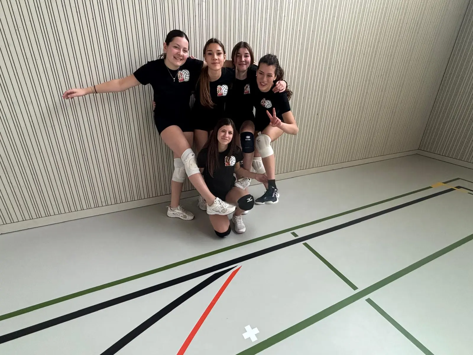 Cinque ragazze in maglietta nera sorridono mentre posano in gruppo. Due di loro sono in piedi sopra le altre tre, che si trovano in ginocchio e sedute. Sono in un ambiente sportivo, con linee di campo visibili sul pavimento.
