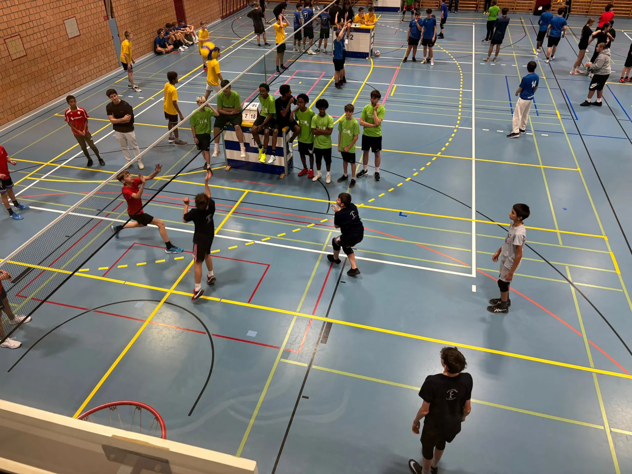 Un palazzetto sportivo affollato con diversi gruppi di ragazzi che giocano a badminton. Alcuni indossano magliette di colori diversi, mentre altri si preparano a colpire il volano. La scena mostra anche le linee del campo e attrezzature sportive