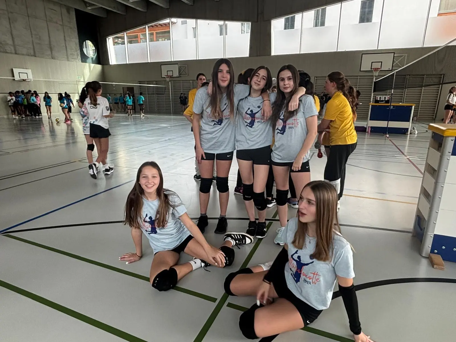Gruppo di giovani atlete di pallavolo in una palestra. Sei ragazze indossano magliette grigie e pantaloni sportivi, sorridendo e posando per la foto. Alcune persone si allenano sullo sfondo.