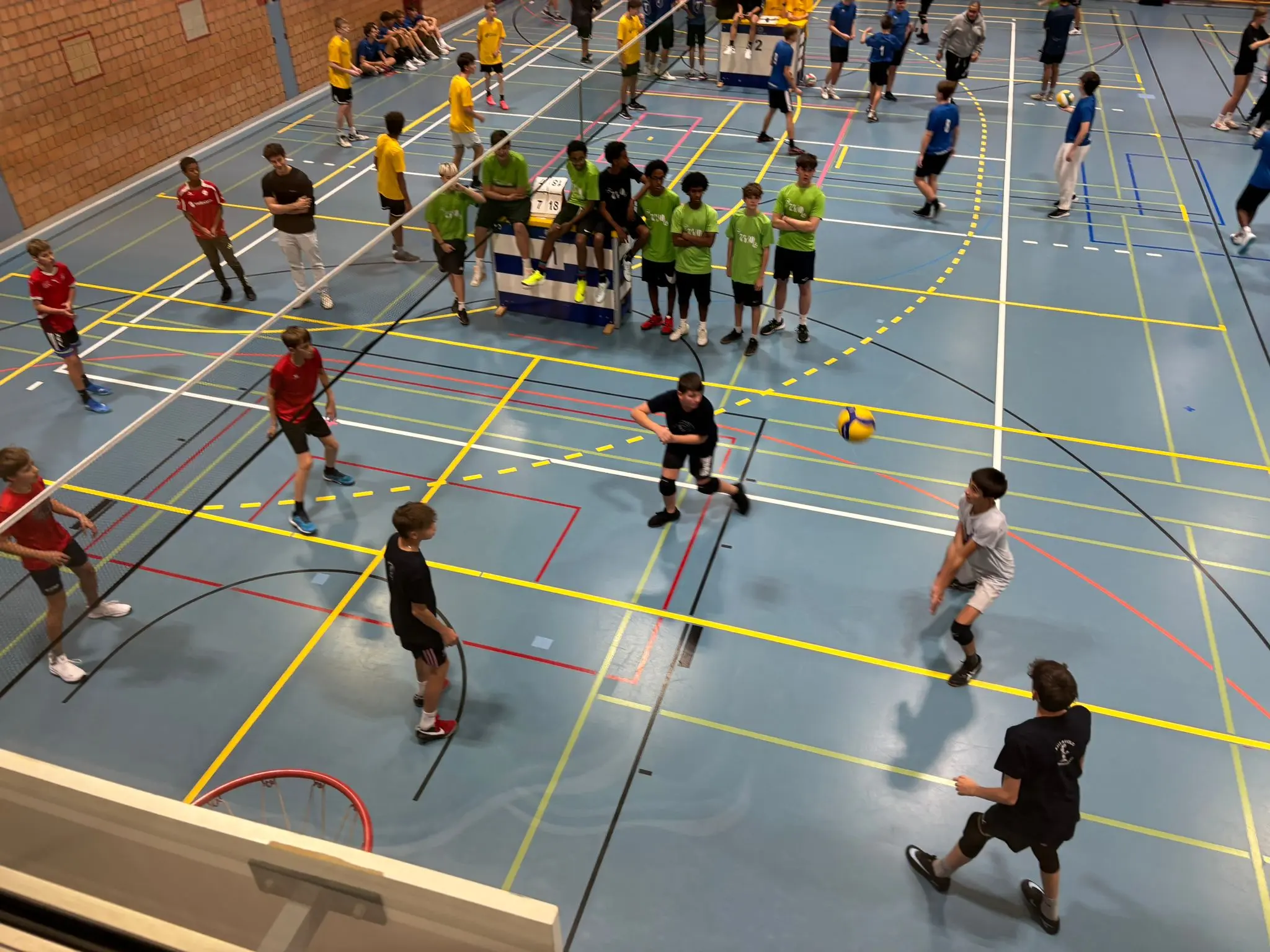 Giovani atleti stanno giocando a pallavolo in un campo sportivo. Ci sono diverse squadre di ragazzi con magliette di colori vari che si preparano e si muovono sul terreno.