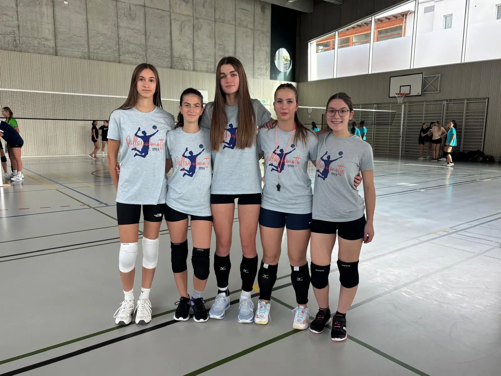 Cinque ragazze in maglietta grigia e pantaloni corti da volley, sorridono in un palazzetto. Alcune indossano ginocchiere e tutti sono in una posizione di gruppo. Sullo sfondo si vedono altre persone che si allenano.