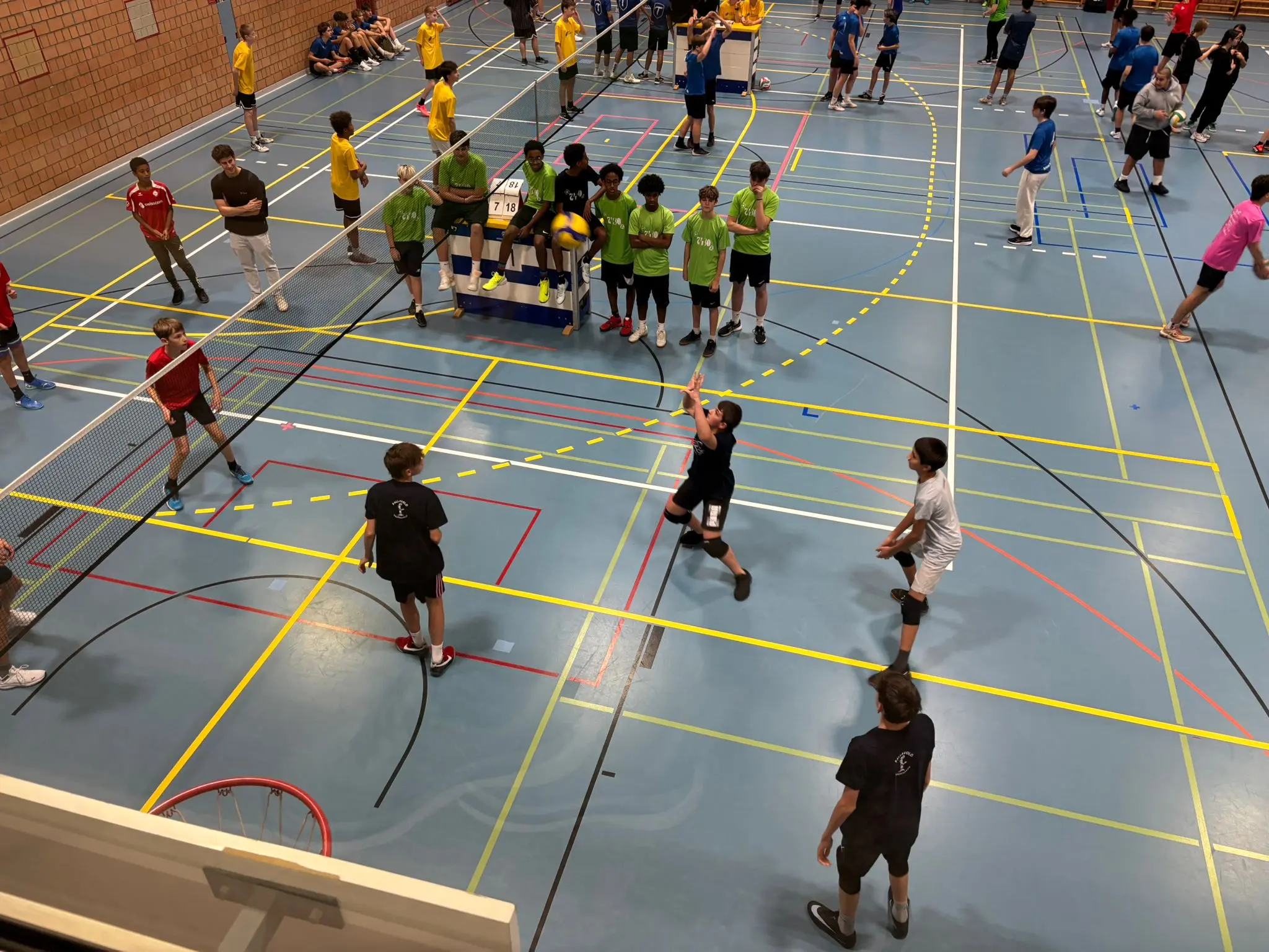 Un gruppo di ragazzi gioca a badminton in un campo sportivo. Sono visibili diverse squadre con magliette colorate, come giallo, verde e blu. Alcuni ragazzi stanno colpendo il volante mentre altri osservano. Il pavimento è segnato con linee color