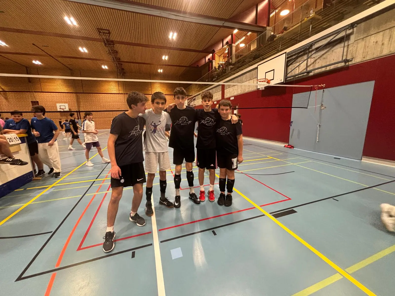 Cinque ragazzi in maglietta nera si abbracciano in un campo di pallavolo al coperto. In sfondo si vedono altri giovani giocatori e attrezzature sportive.