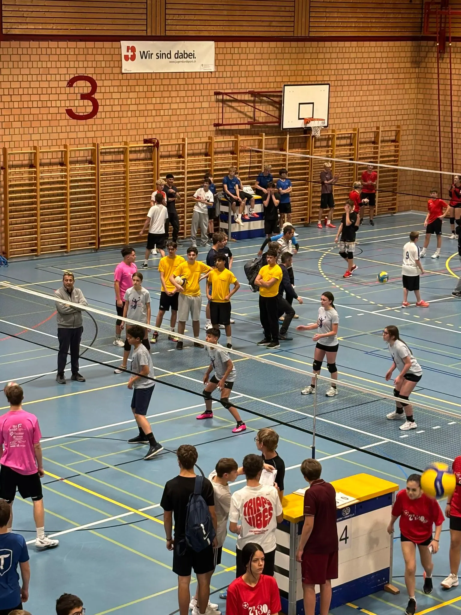 Un gruppo di ragazzi e ragazze si allenano in una palestra di badminton. Ci sono diversi team, alcuni indossano magliette gialle e altri grigie. Alcuni atleti si preparano a ricevere la palla mentre altri osservano.
