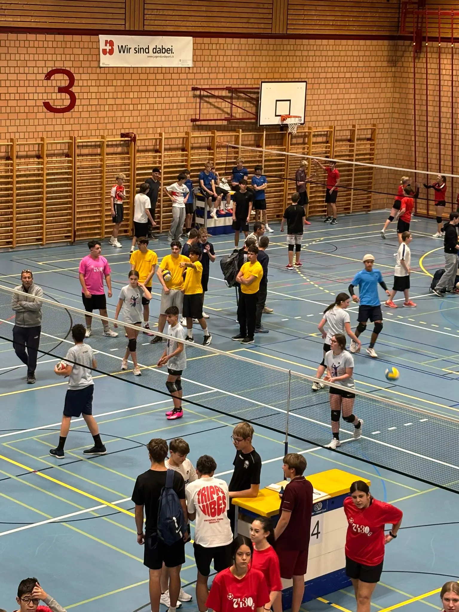Un gruppo di giovani atleti si allena in un pallone ginnico, divisi in diverse squadre. Alcuni indossano magliette colorate, mentre altri praticano esercizi di riscaldamento. Circondati da attrezzature sportive e reti.