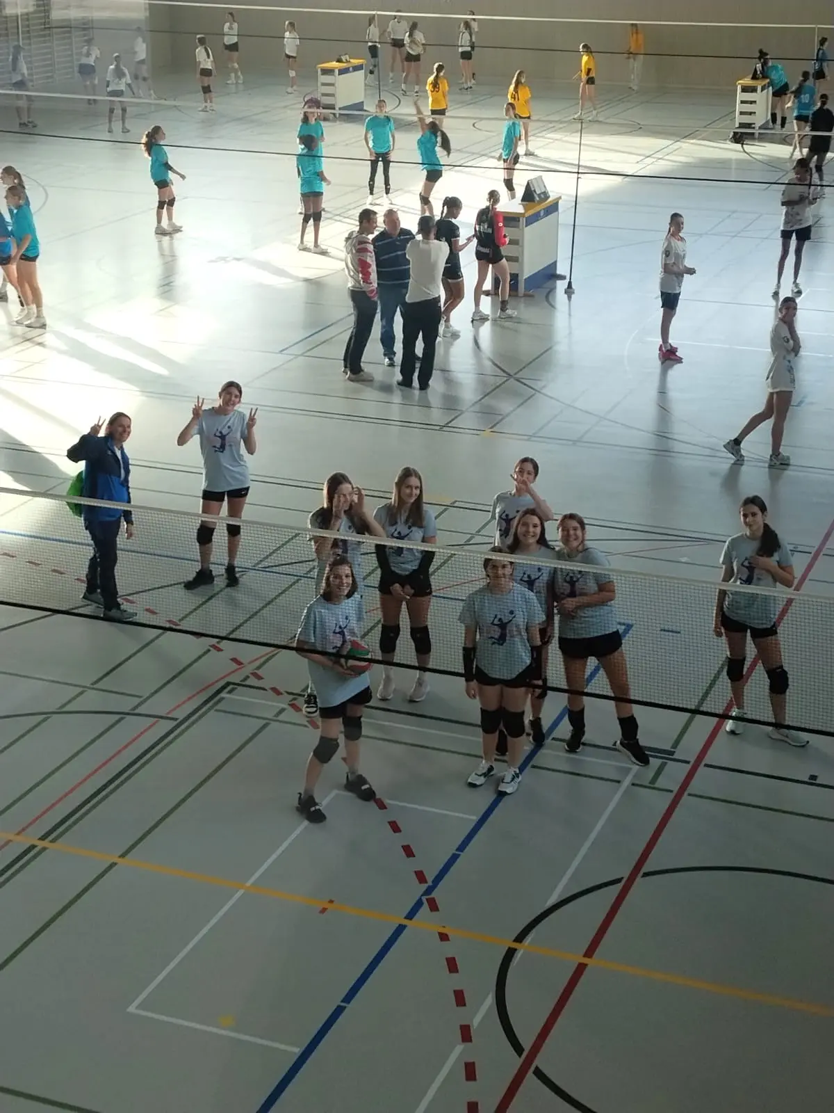 Un gruppo di ragazze in maglietta grigia sorride e fa segni di pace mentre posano davanti a una rete da pallavolo. Sullo sfondo, altre persone sono impegnate in diverse attività sportive in un ampio campo.