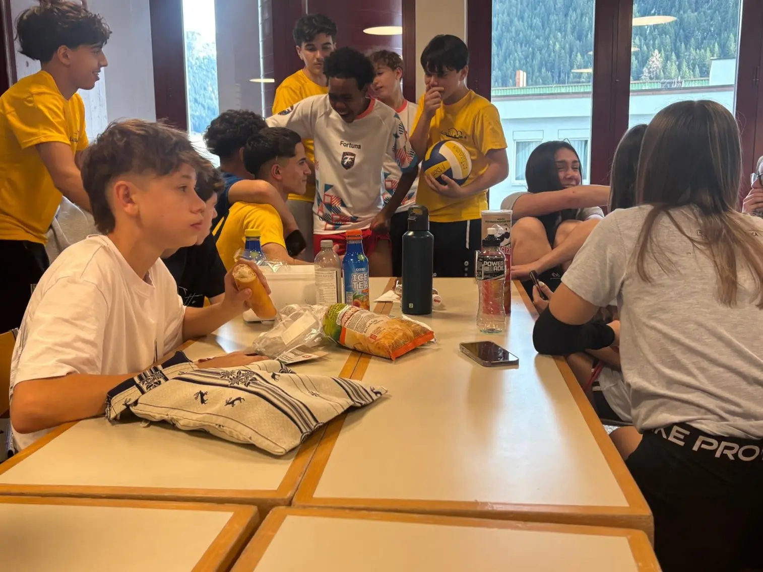 Un gruppo di ragazzi in maglietta gialla e bianca conversa e gioca attorno a un tavolo. Alcuni si abbracciano, mentre altri mangiano snack e bevono da bottiglie d'acqua. Sullo sfondo si intravedono montagne.