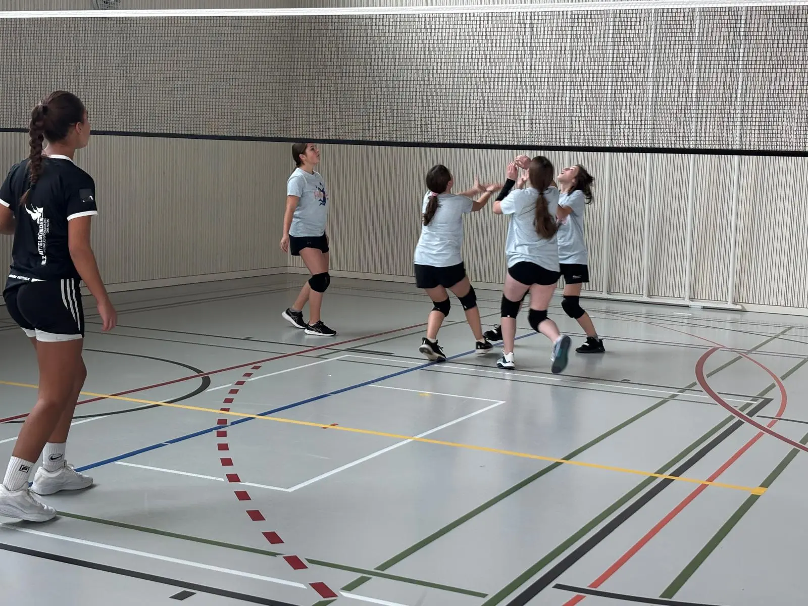 Un gruppo di ragazze gioca a pallavolo in una palestra. Alcune si preparano a ricevere la palla, mentre una ragazza sul lato sinistro osserva. Le atlete indossano magliette azzurre e pantaloni corti.