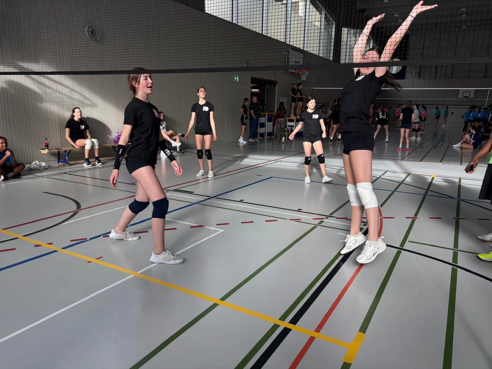 Giovani atlete di pallavolo in un momento di gioco in palestra, con una giocatrice che salta per colpire la palla mentre le altre osservano. La scena mostra un ambiente energetico e competitivo.