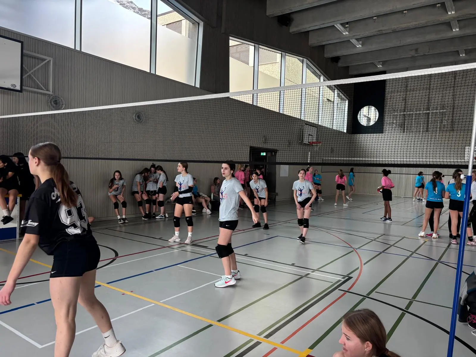 Un gruppo di ragazze si allena a pallavolo in un palazzetto moderno. Alcune giocano attivamente, mentre altre osservano o si preparano a entrare in campo. La luce naturale entra attraverso le finestre.