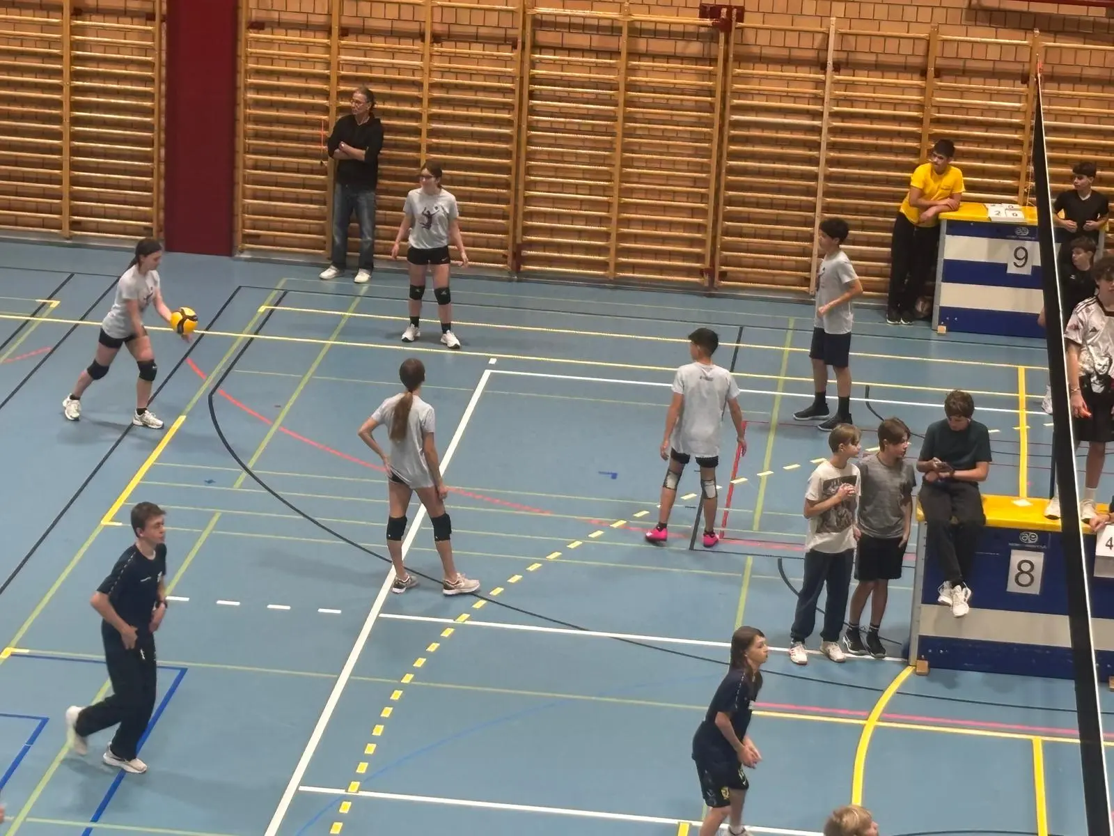 Una palestra con diverse persone che giocano a pallavolo. Quattro giocatori sono in campo, mentre altri si trovano a bordo. Alcuni osservano e discutono.