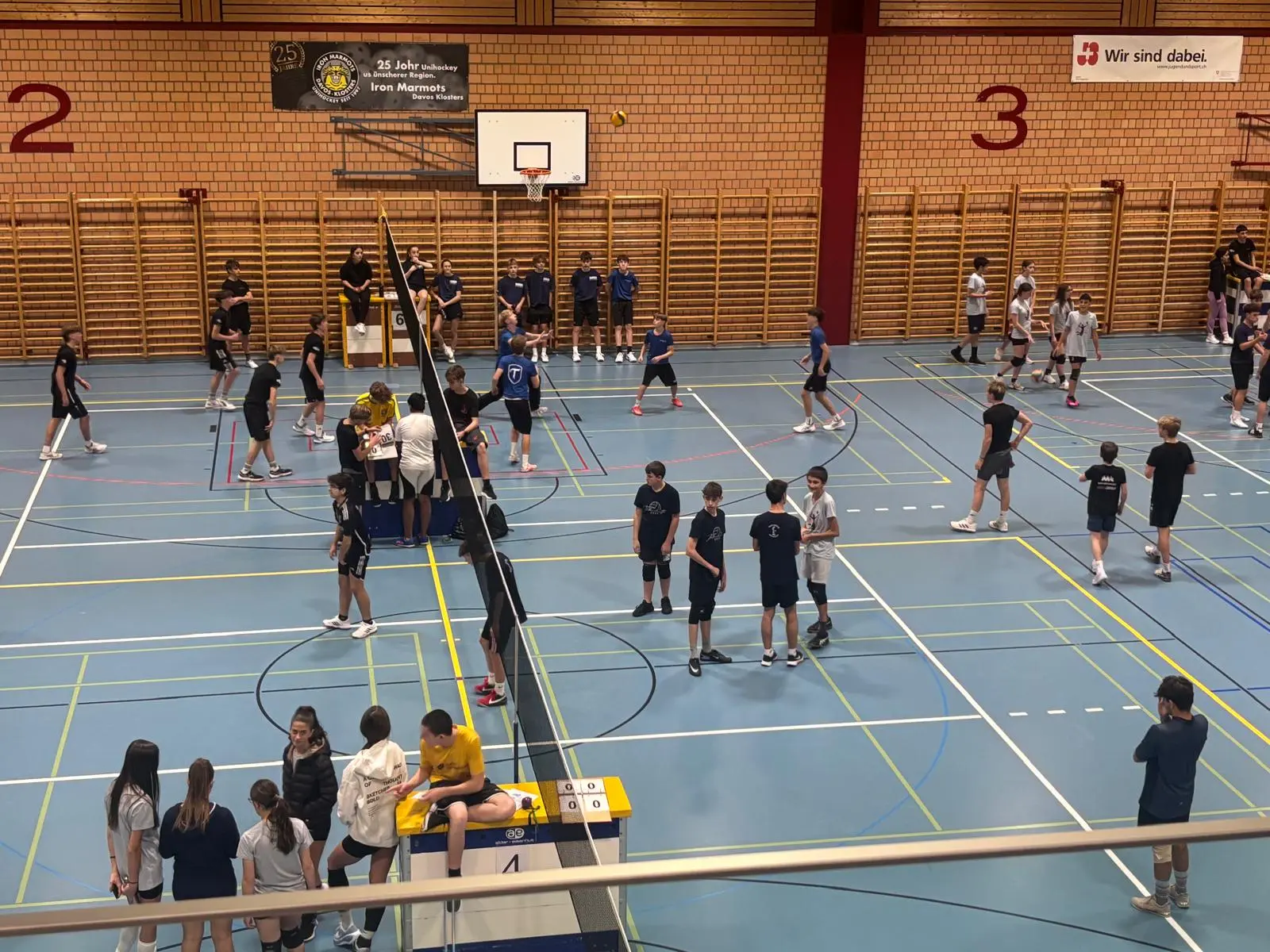 Un palazzetto dello sport affollato con ragazzi e ragazze che giocano a pallavolo. Ci sono due squadre in campo e altre persone che si preparano a giocare o che osservano. La rete e il tabellone sono ben visibili.