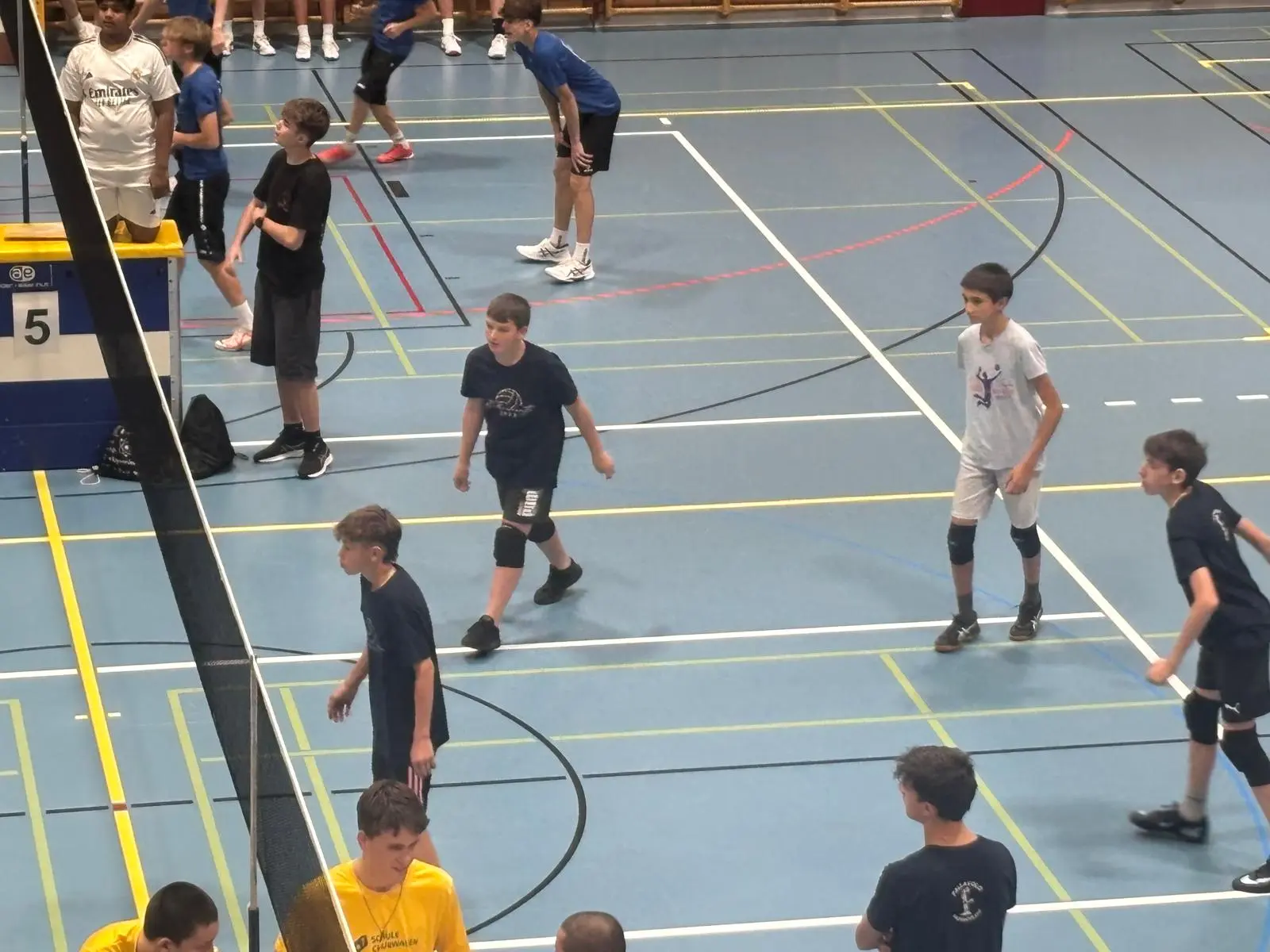 Gruppo di ragazzi in campo di pallavolo, pronti per iniziare una partita. Indossano magliette e pantaloni sportivi, alcuni sono in posizione di attesa, mentre altri si preparano. Dietro di loro si intravedono altri giocatori e il campo è