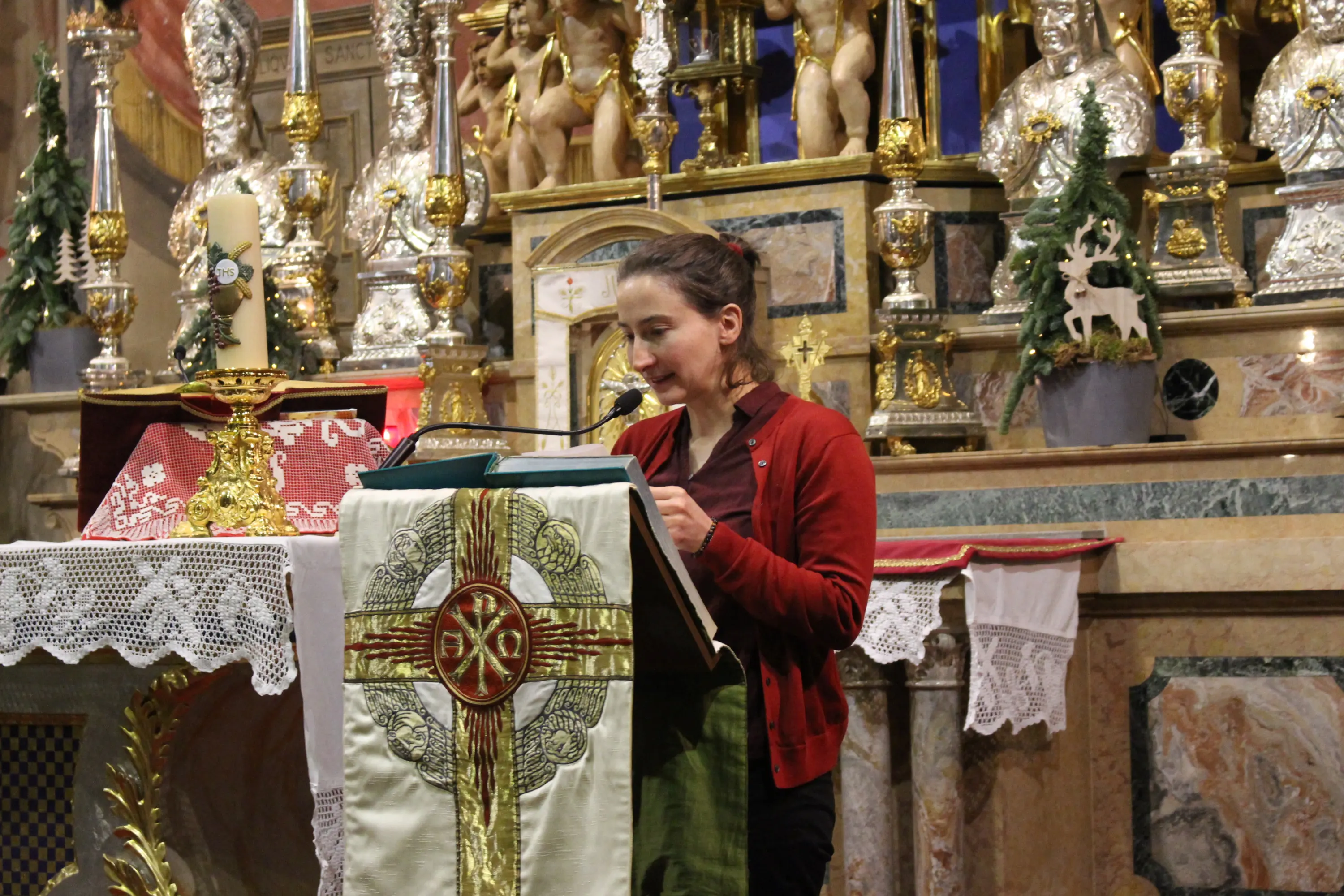 Una giovane donna legge da un leggìo in una chiesa, con un altare decorato sullo sfondo. L'altare è ornato con statui, candele e dr