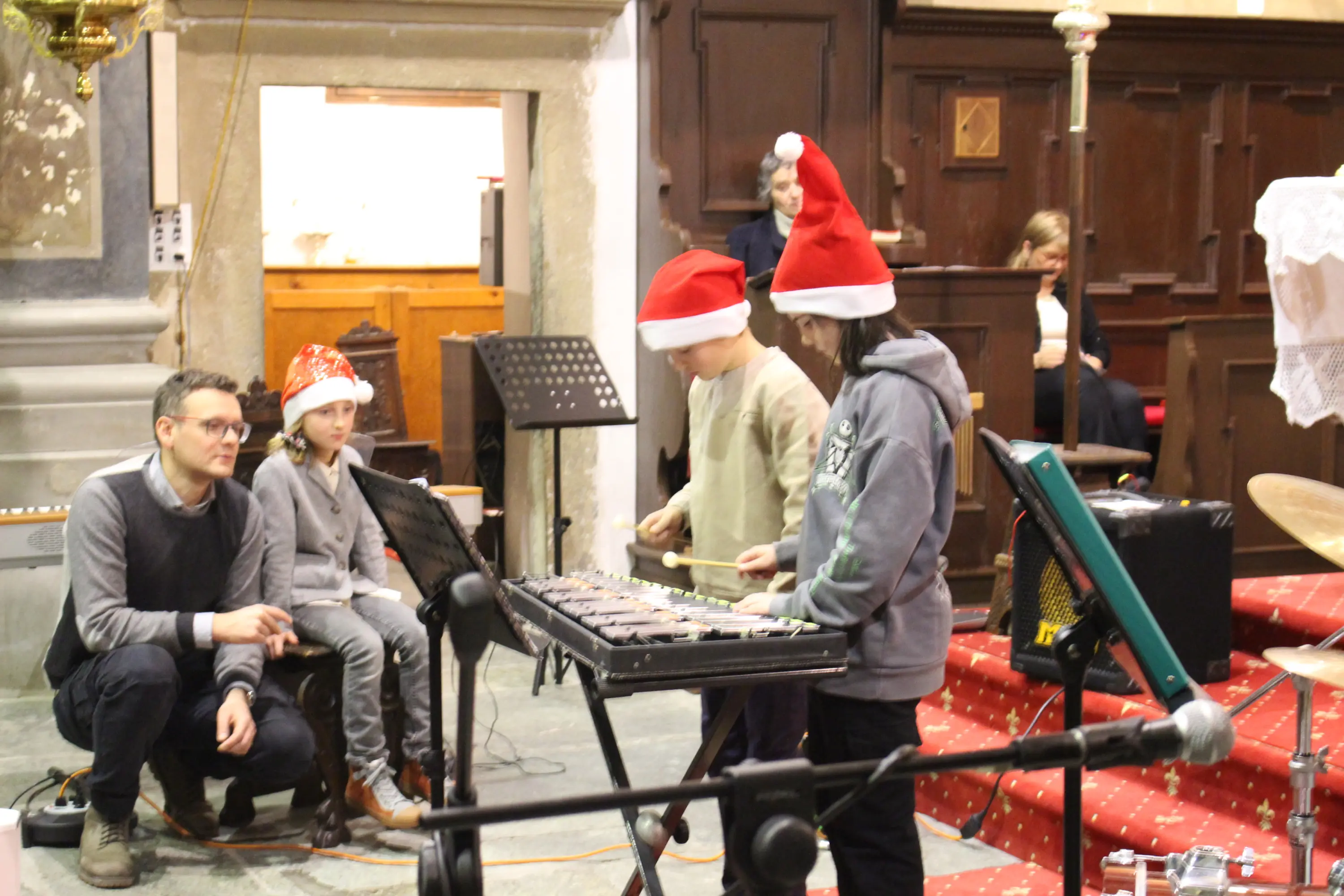 Un gruppo di bambini suona uno xilofono in una chiesa, indossando cappelli di Natale. Un adulto li osserva e si sta preparando a offrir loro supporto. Sullo