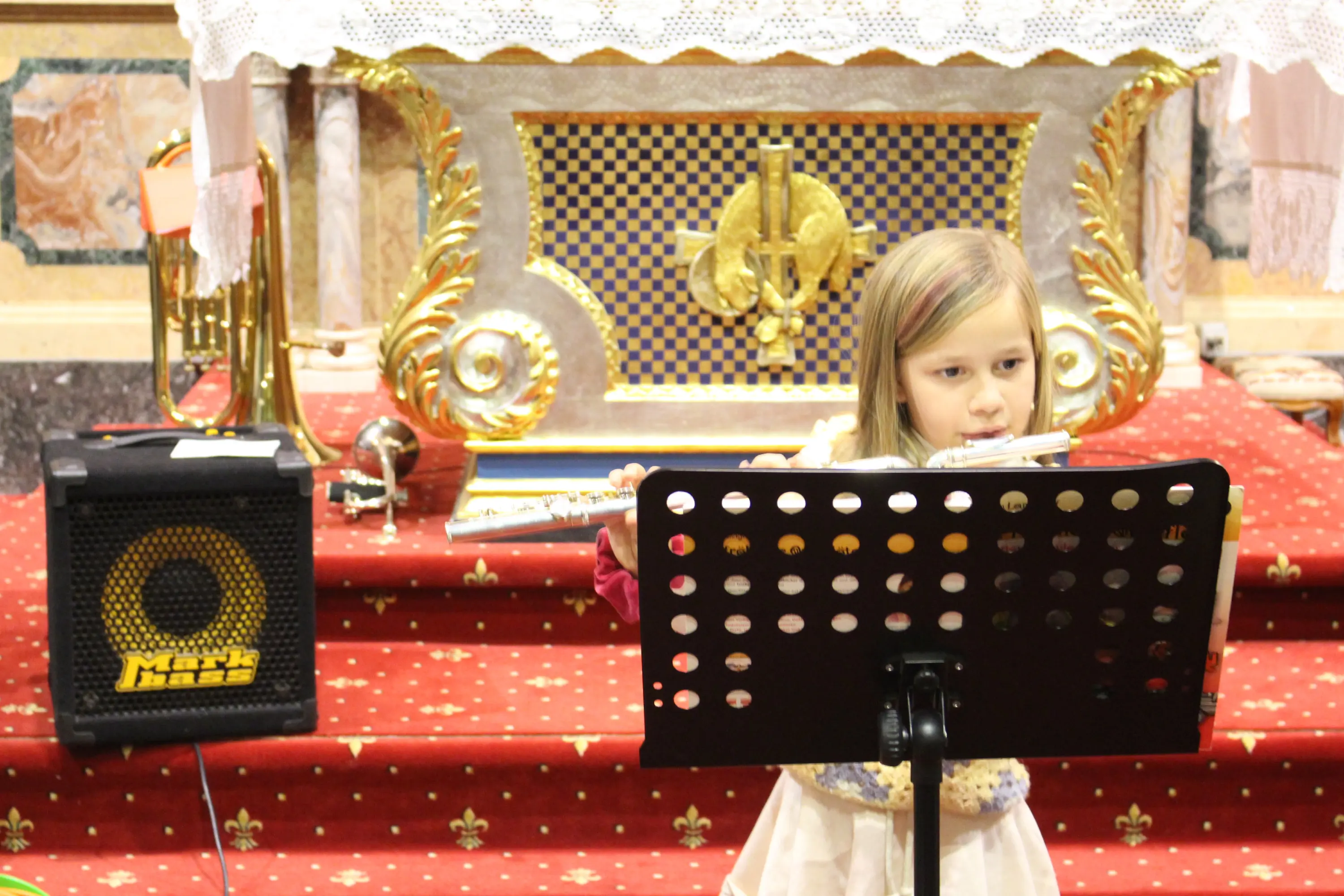 Una bambina suona il flauto davanti a un leggio, all'interno di una chiesa. Sullo sfondo si vede un altare decorato e un amplificatore.