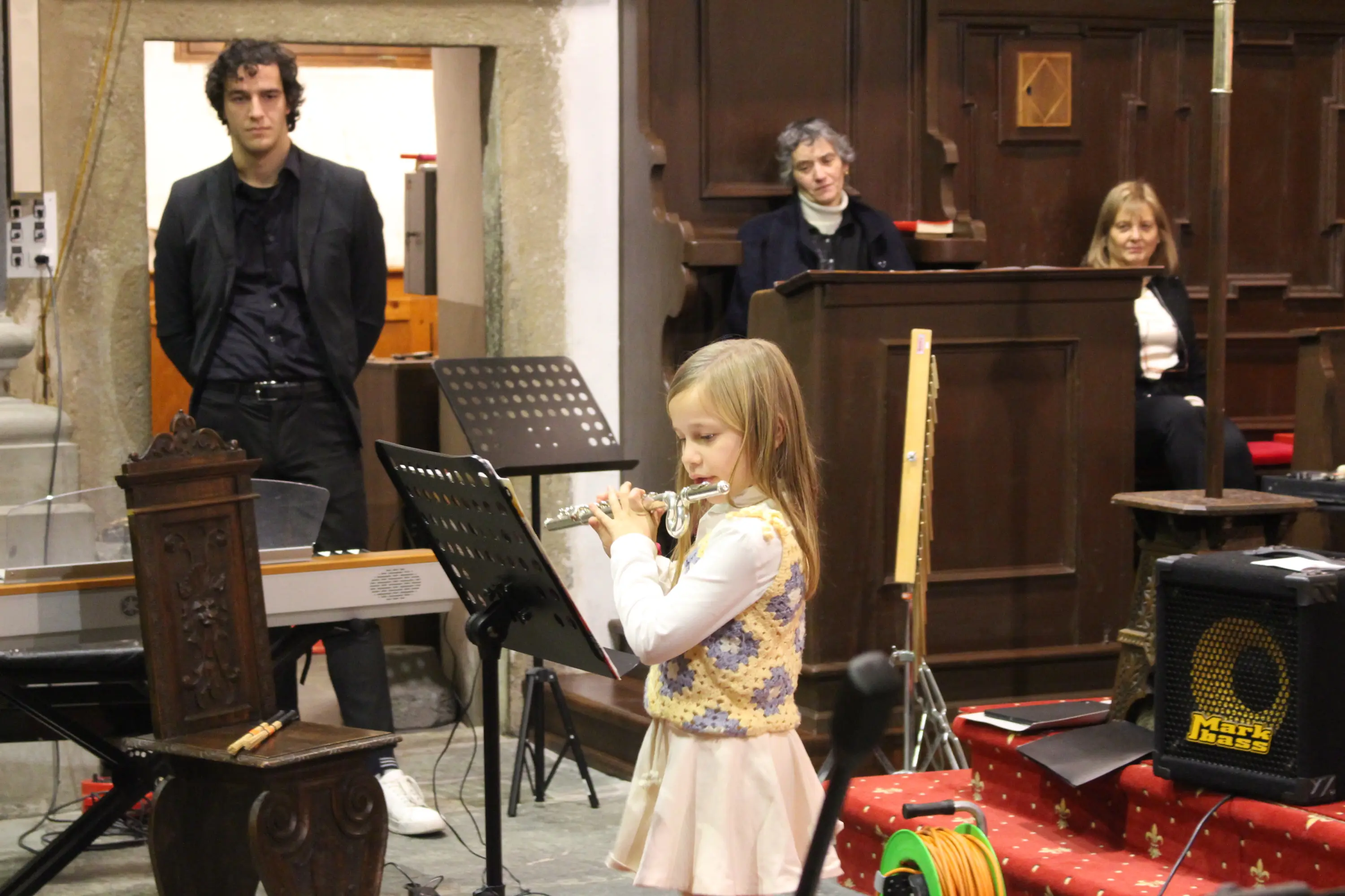 Una bambina suona il flauto in un ambiente di concerto. Tre adulti la osservano da dietro, in una sala con pareti in legno e attrezzature musicali.