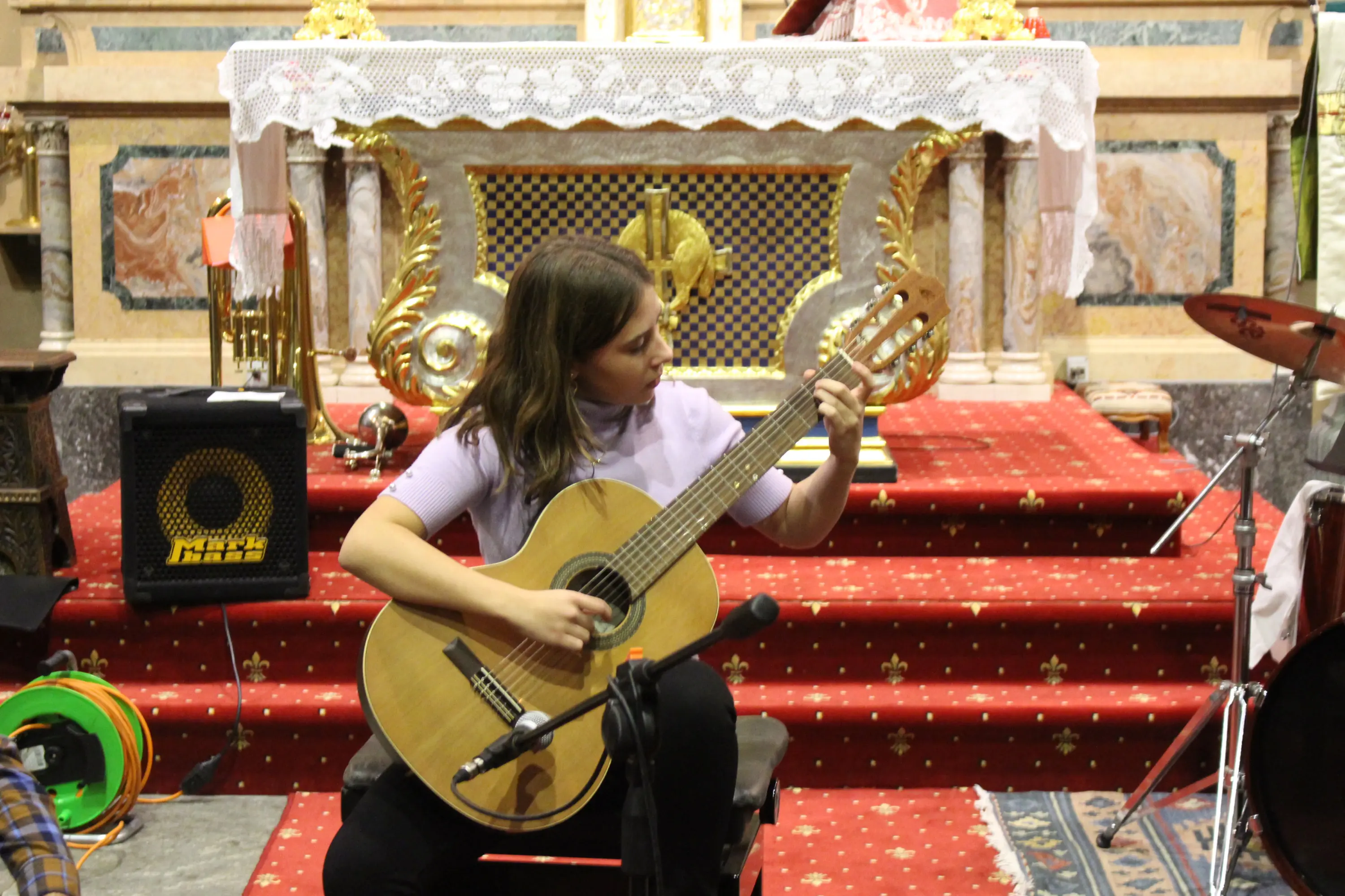 Una musicista suona la chitarra in un ambiente accogliente, con un altar decorato sullo sfondo. Il pavimento è coperto da un tappeto rosso e ci