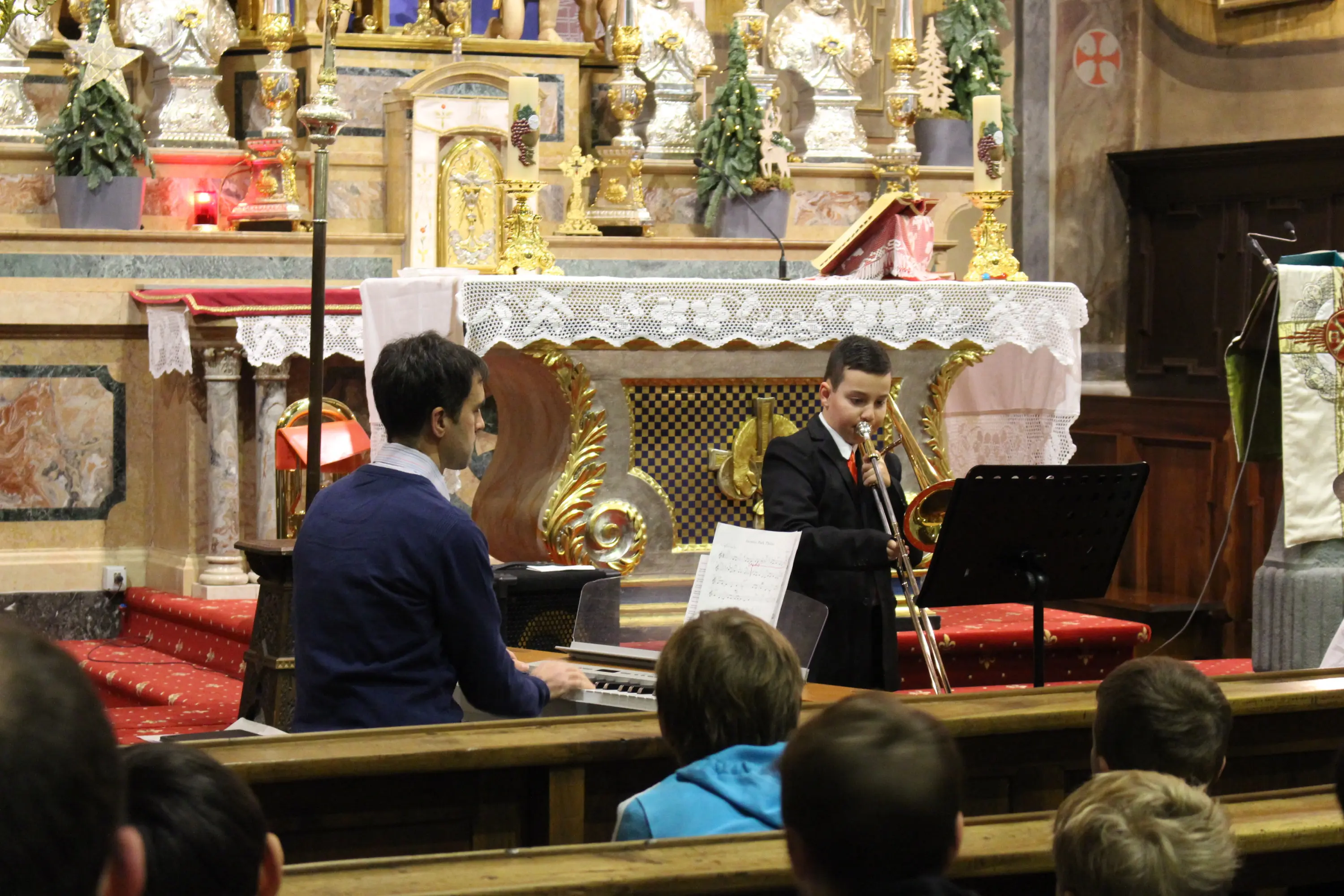 Un bambino in abito elegante suona il trombone mentre un adulto lo accompagna al pianoforte in una chiesa decorata. In primo piano, un gruppo di ragazzi ascolta attentamente