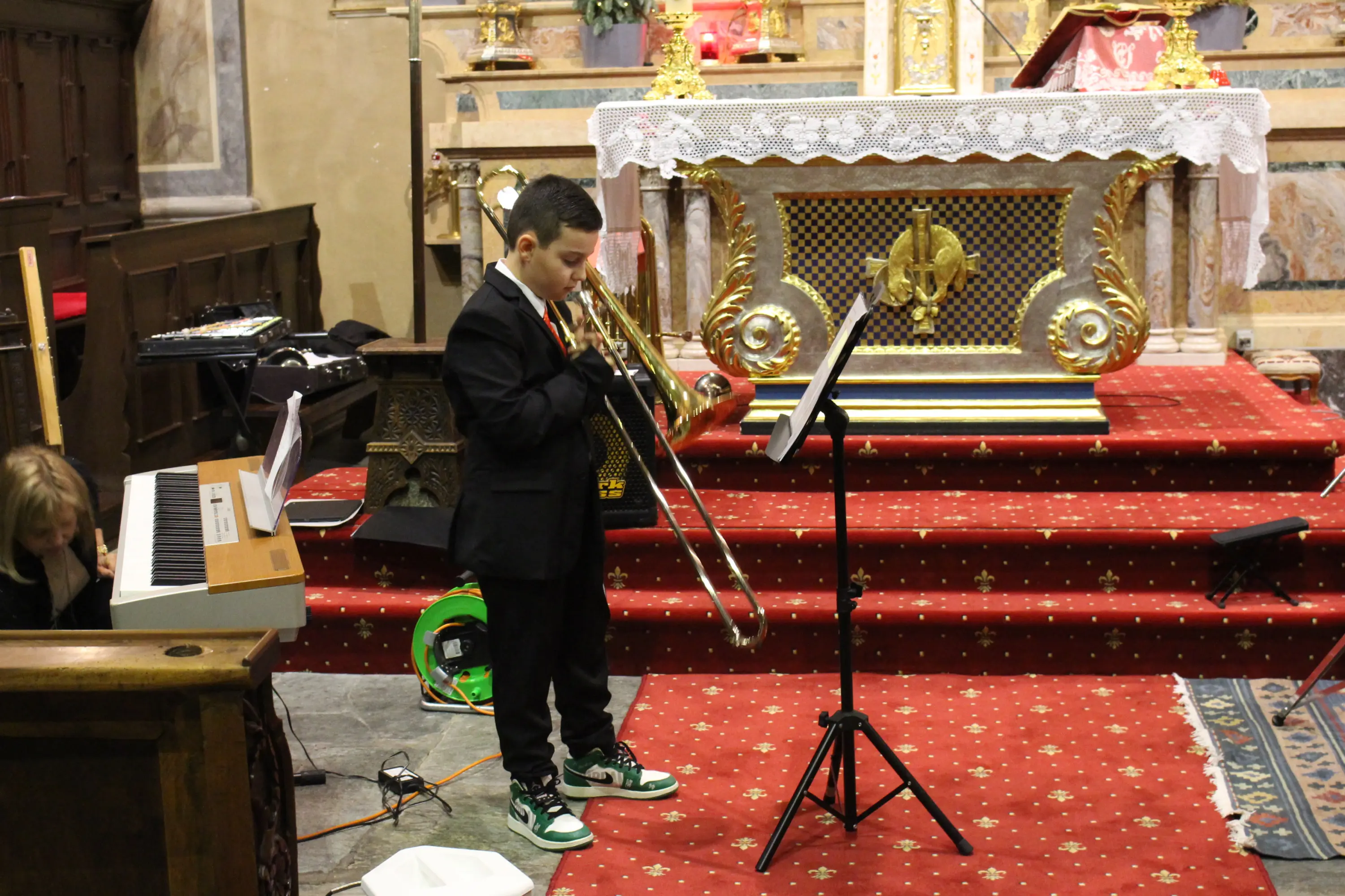 Un ragazzo vestito elegante suona il trombone in una chiesa, mentre un pianoforte e alcune attrezzature musicali sono visibili sul lato. L'altare è decorato
