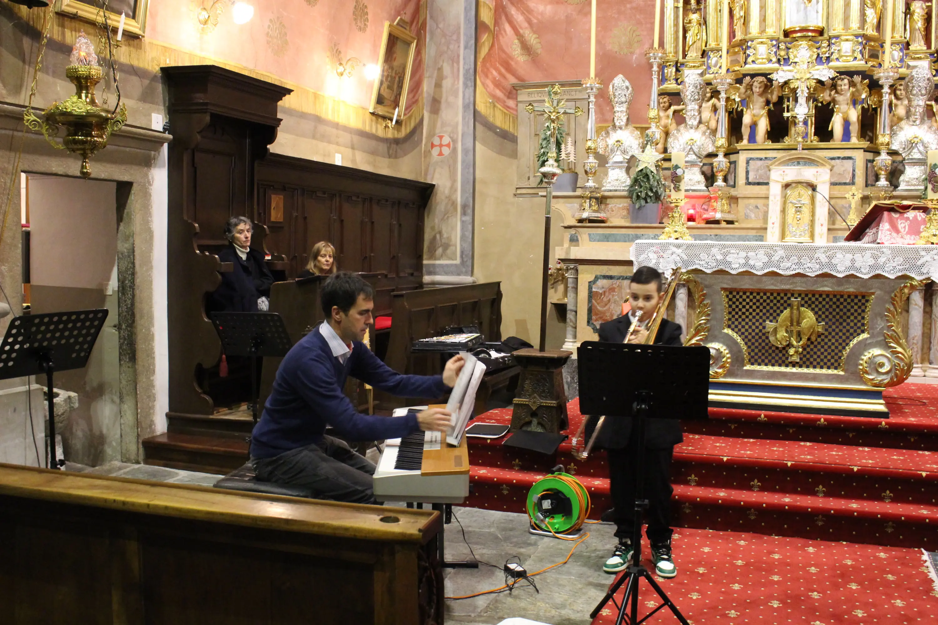 Un uomo suona il pianoforte mentre un ragazzo in giacca e cravatta suona il flauto davanti a un altare decorato. Alcuni spettatori sono seduti sul